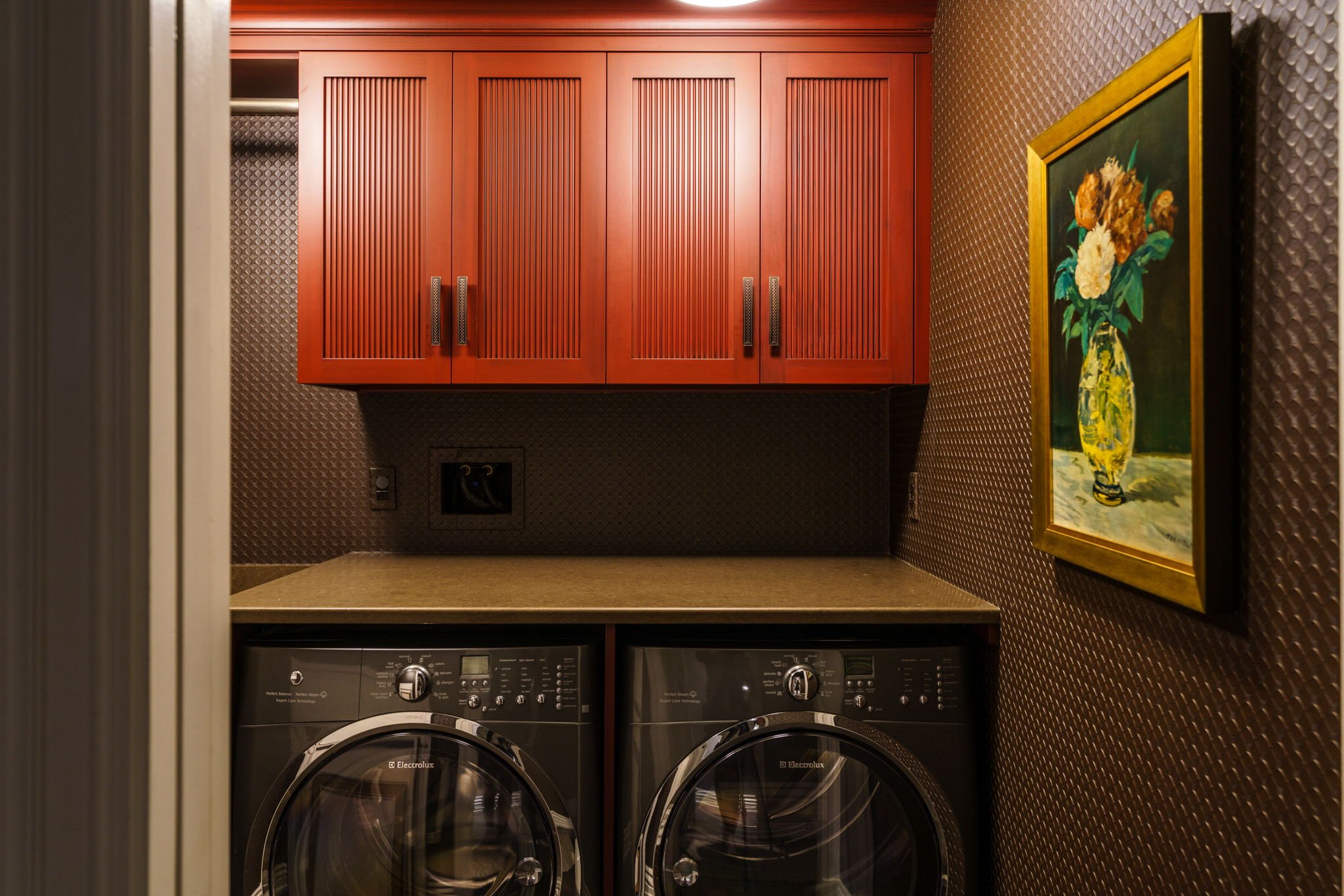 A laundry area with two front-loading Electrolux washers stacked side by side, a wooden cabinet with four doors above, and a framed floral still life painting on the patterned wall.