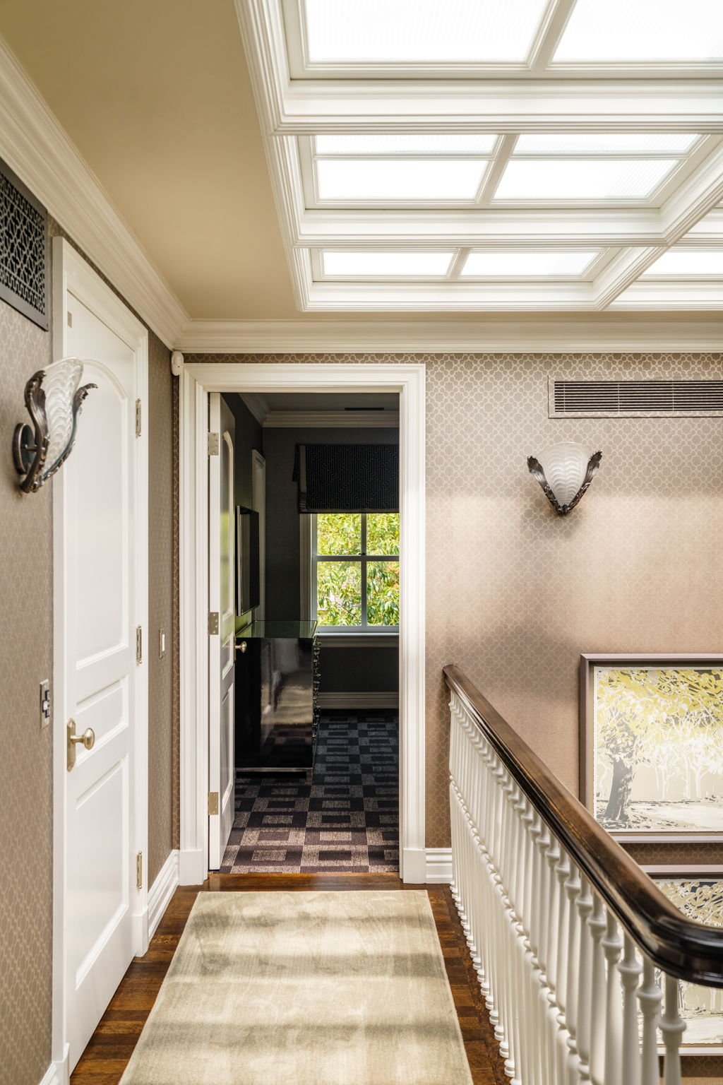 Hallway with patterned wallpaper, white doors, a beige rug, and wall sconces, leading to a room with a window showing green foliage outside.