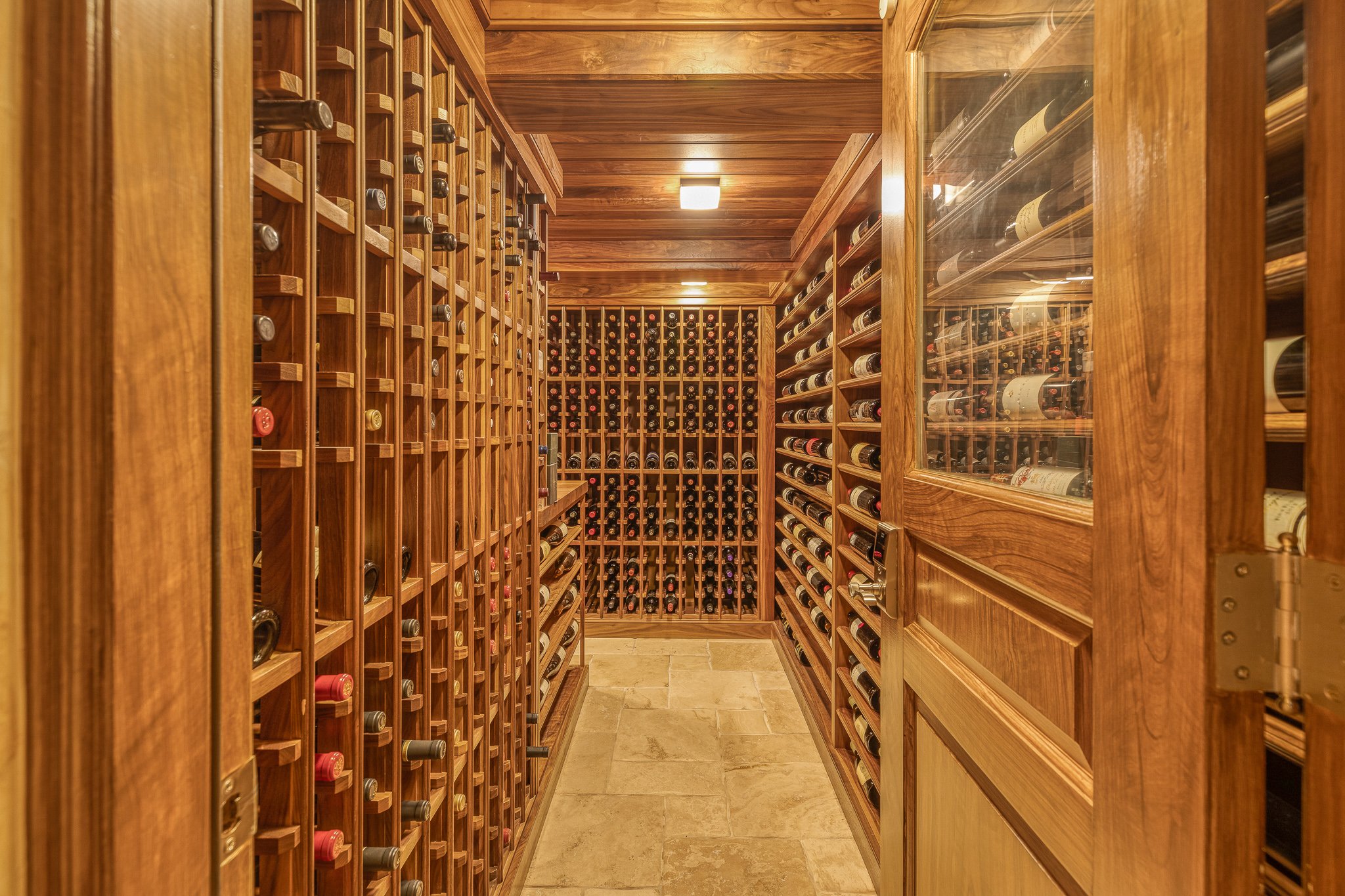 A wine cellar with wooden shelves filled with wine bottles, a wooden ceiling, and a stone floor.