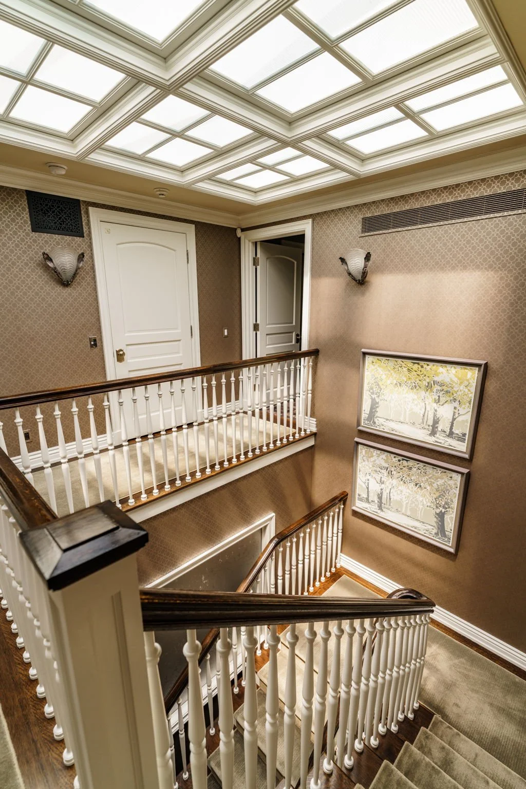 Interior view of a well-lit staircase landing with beige wallpaper, white doors, framed artwork, and wall sconces.
