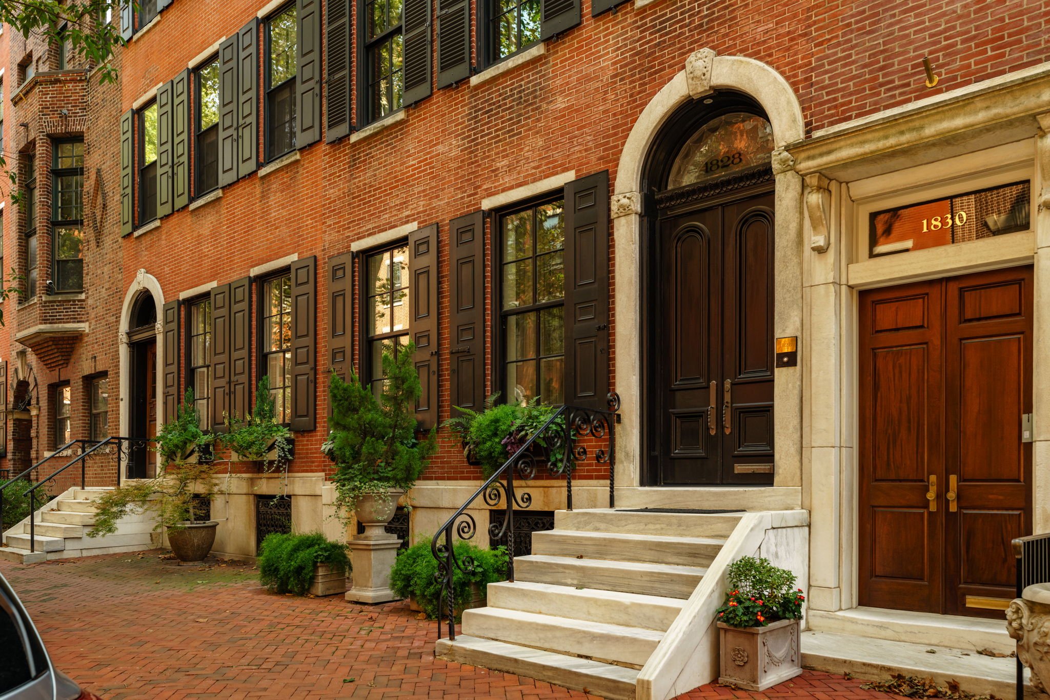 Brick townhouse with black shutters, arched front door with stone surround, and stairs leading up to the entrance, with potted plants on the steps and sidewalk.