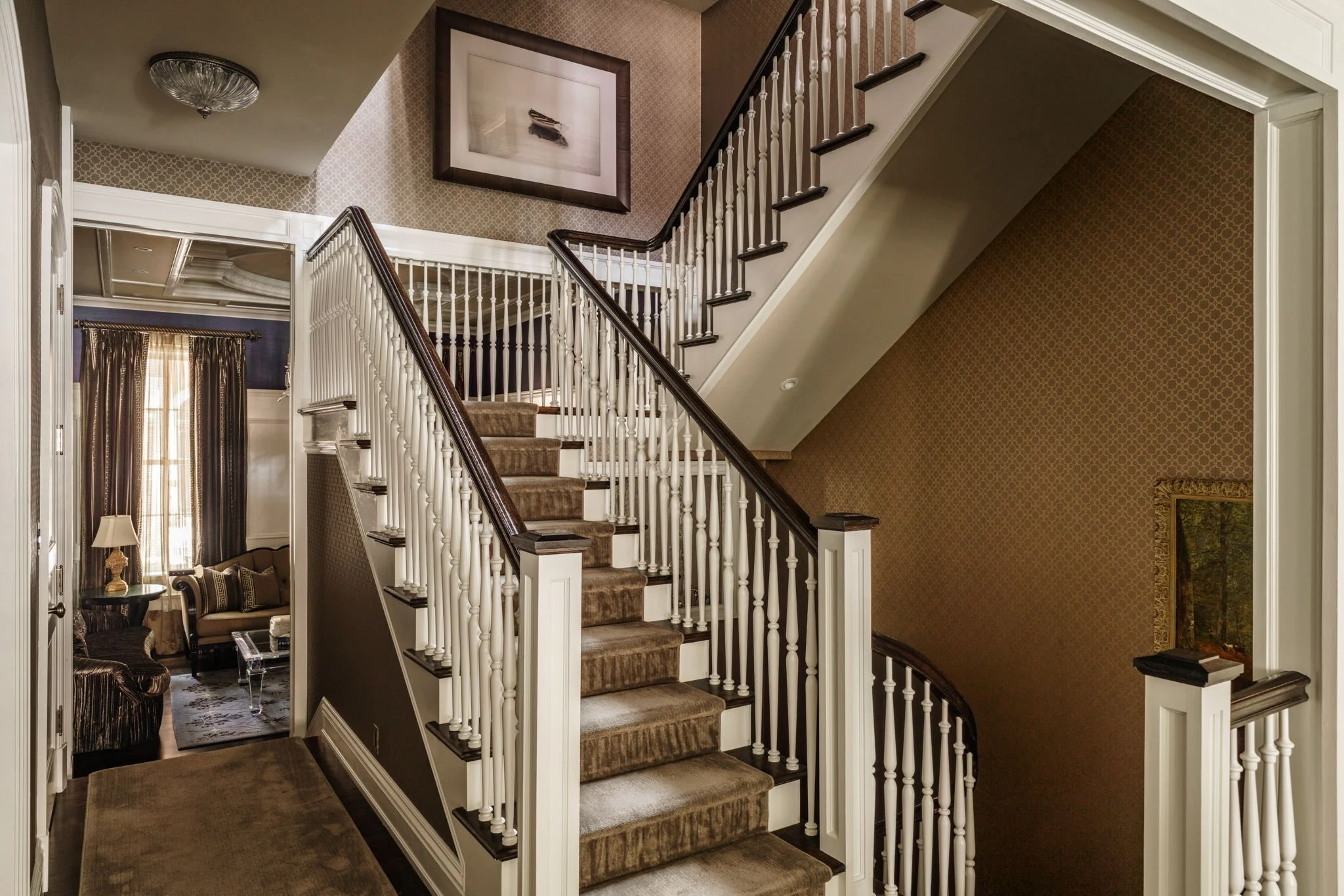 Staircase in an elegant home with beige carpeting, white spindles, and dark wood handrails, leading to upper level with framed artwork on the wall.