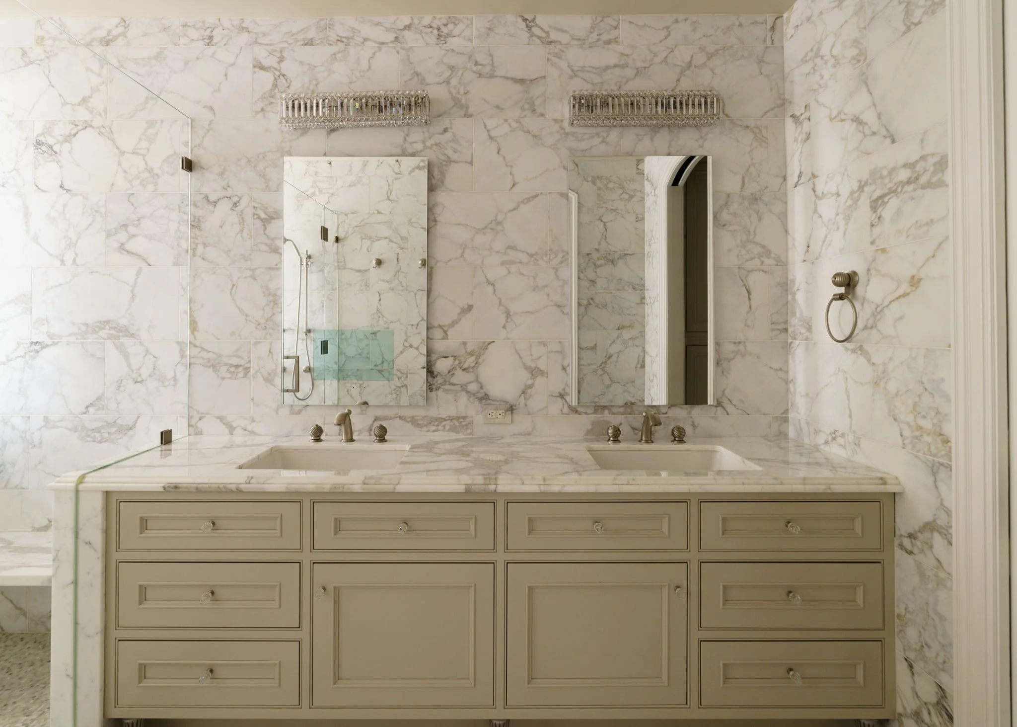 A bathroom with marble walls, two crystal light fixtures, a large mirror, a double sink vanity with drawers, and a glass shower enclosure.