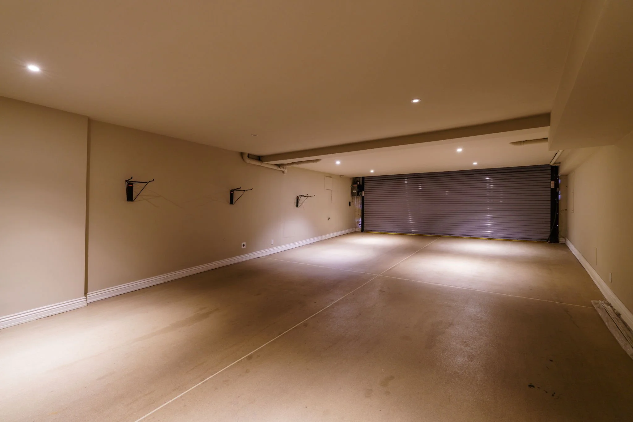Empty garage with beige walls, concrete floor, and a metal roll-up door at the far end. Several wall-mounted light fixtures are visible along the left wall. The garage is well-lit with recessed ceiling lights.