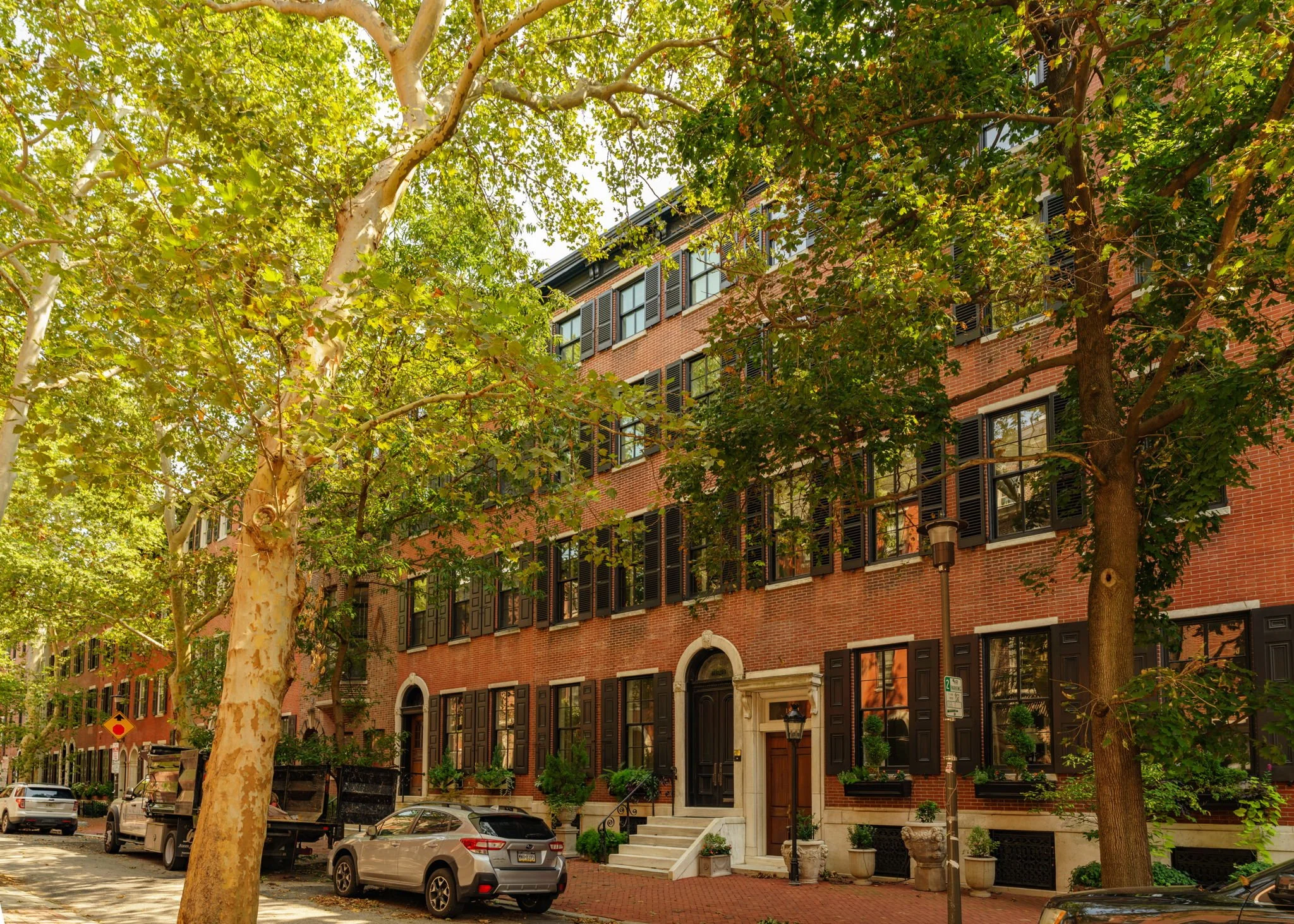 Tree-lined city street with brick townhouses, parked cars, and trees with green leaves