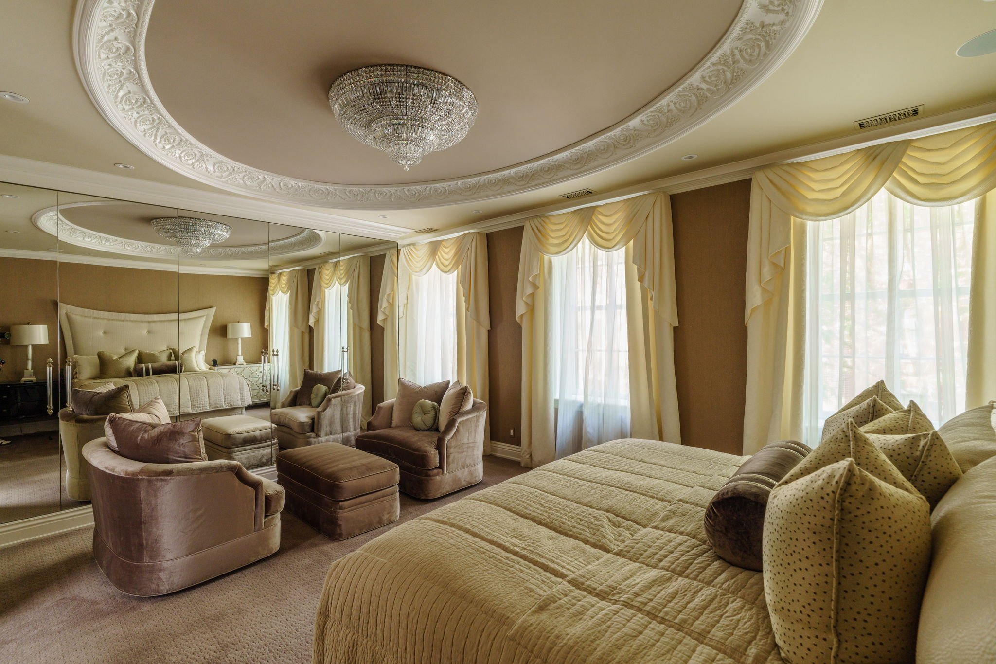 Luxurious bedroom with velvet armchairs, a large bed with patterned bedding, and windows with elegant cream drapes, decorated with ornate crown molding and a crystal chandelier.