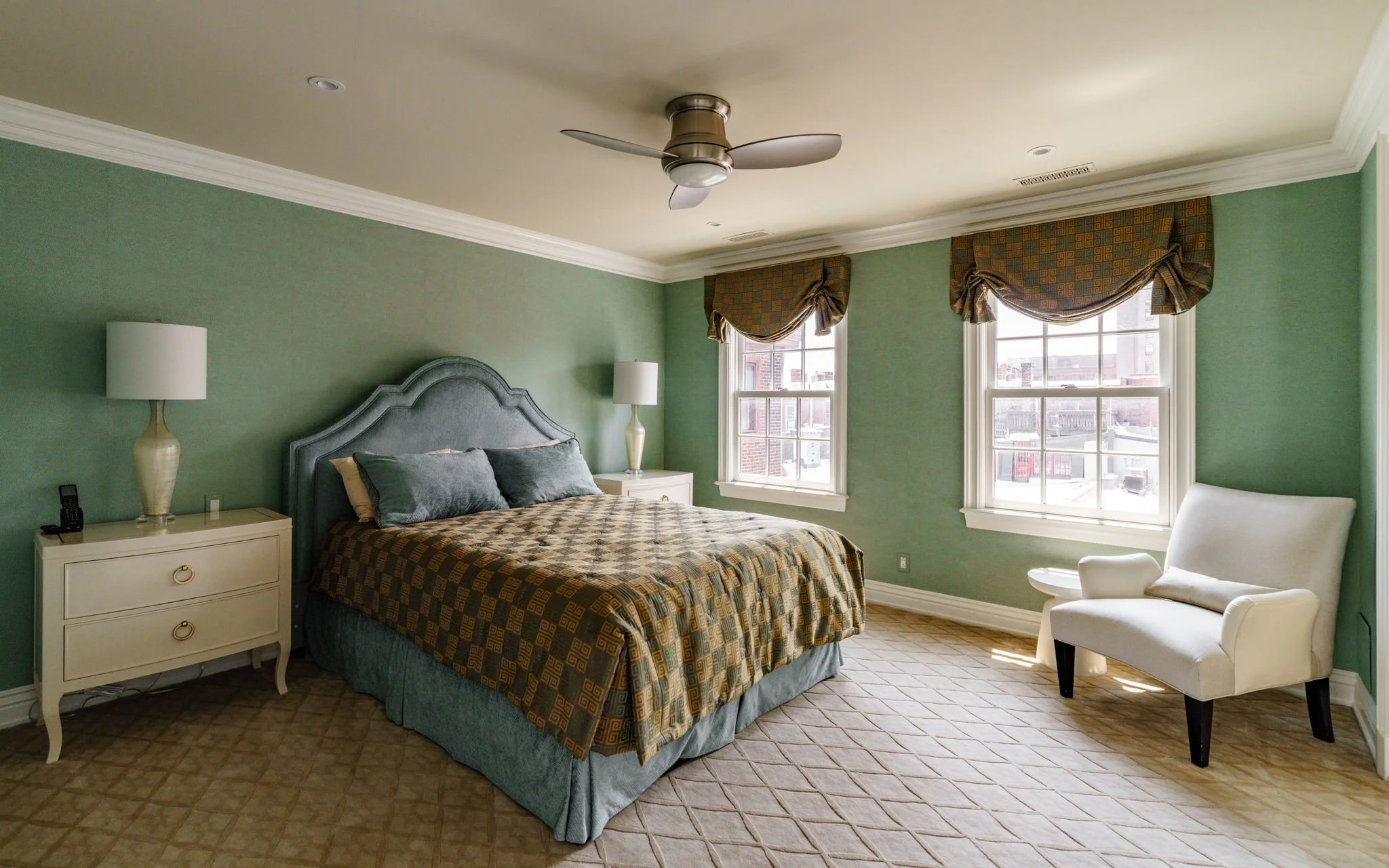A bedroom with green walls and a beige ceiling, featuring a bed with a patterned comforter, two nightstands with lamps, a white armchair, and windows with tie-up valances.