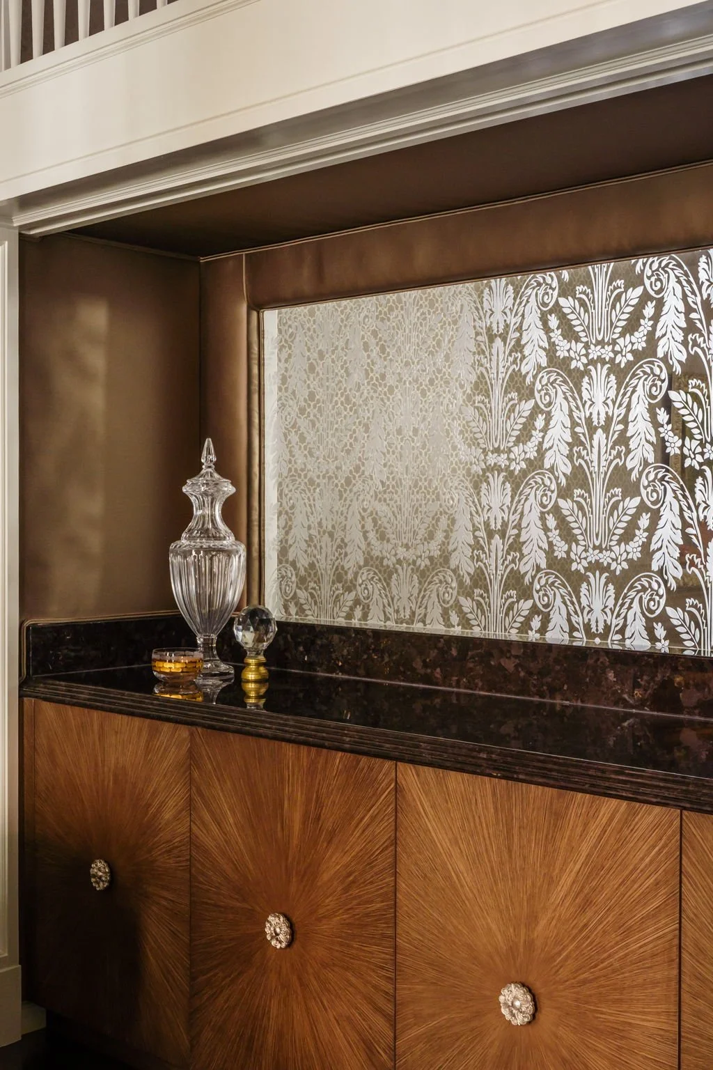 A decorative wooden cabinet with a dark marble top, adorned with a glass decanter and small glass container, against a wallpapered wall with a white floral pattern.