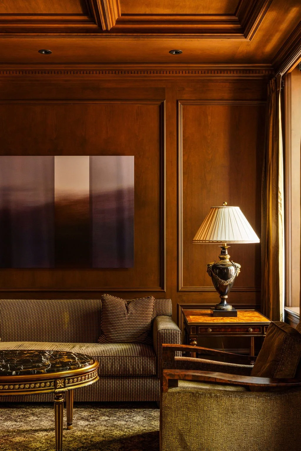 A luxurious living room with wooden paneled walls, a patterned sofa, a marble-topped coffee table, a side table with a decorative lamp, and a large window with curtains.