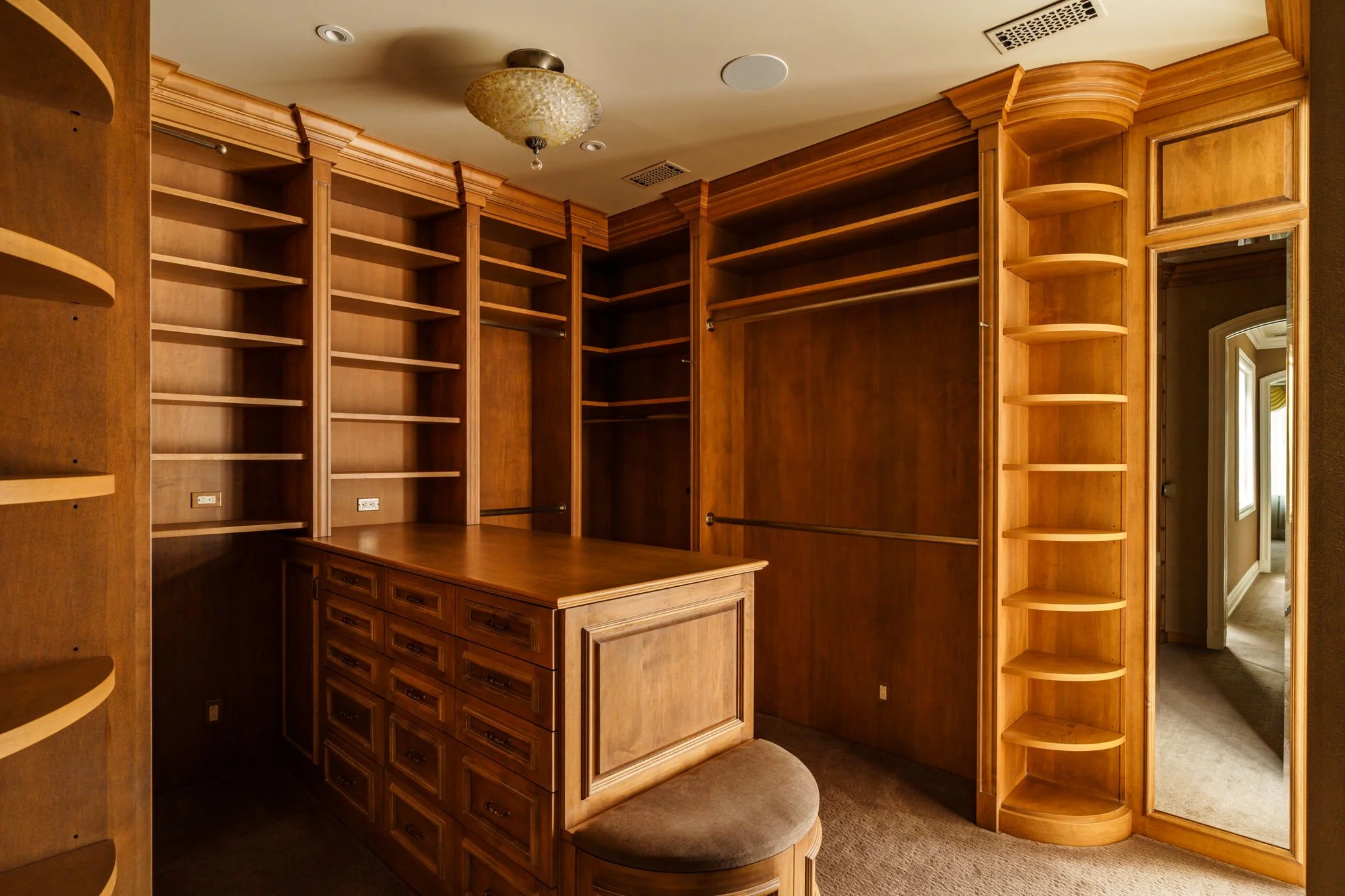 Wooden built-in corner shelves and cabinets in a room, with some panels open revealing empty shelves, a small seating area with a cushioned stool, and a mirror on the right side.