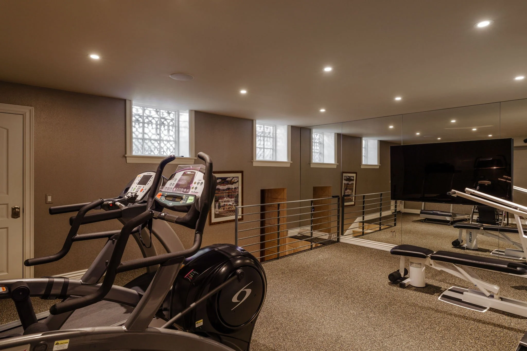 Interior view of a home gym with exercise equipment including treadmills and a weight machine, beige walls, large mirrors, and windows with decorative glass blocks, lit by ceiling lights.