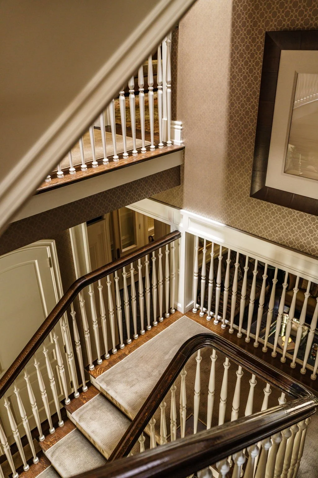 View of a staircase with beige carpeted steps, dark wooden handrails, and white spindles, next to walls with patterned beige wallpaper and framed artwork.