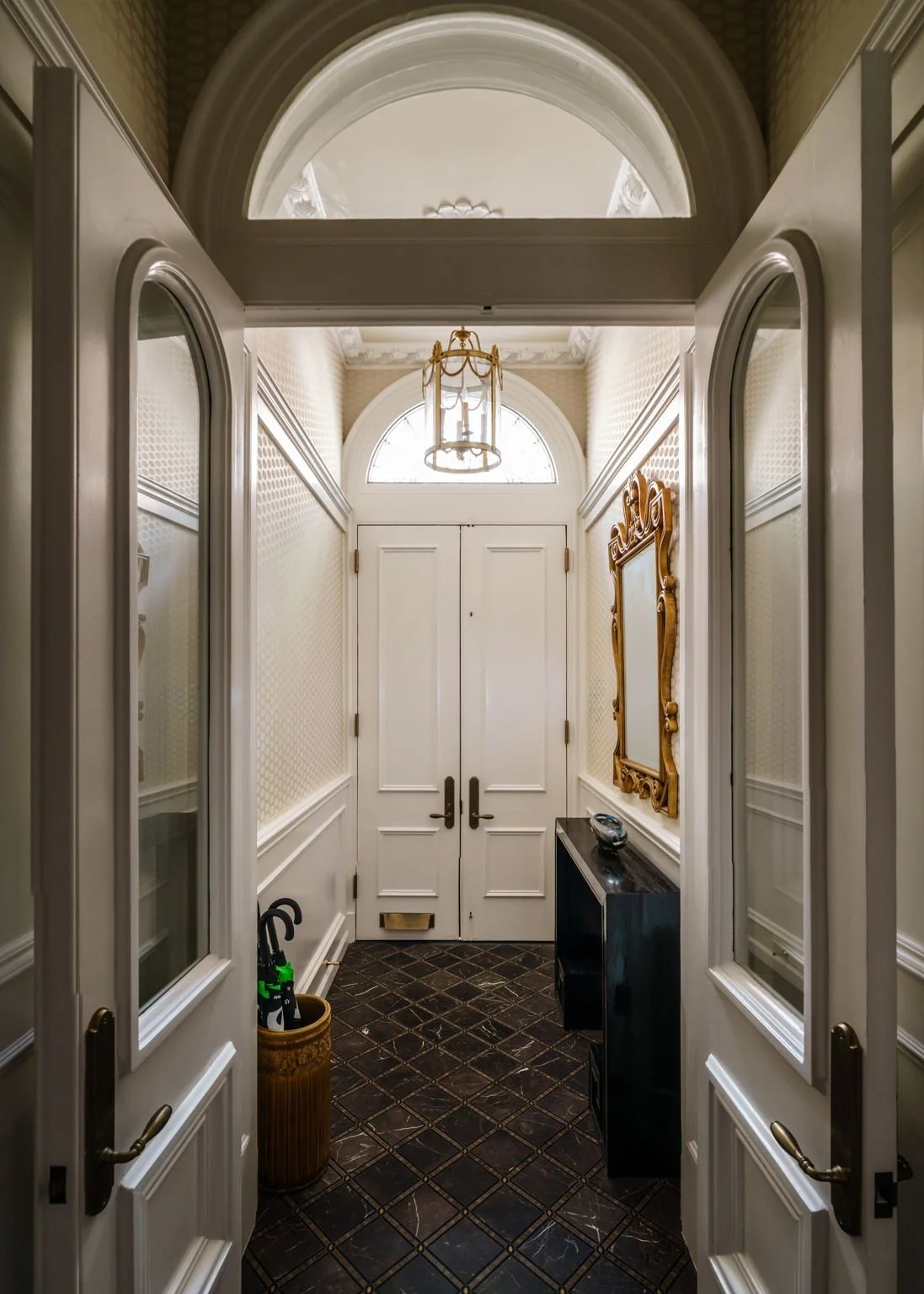 A view of an elegant foyer with double white doors, a black floor with gold accents, a gold framed mirror on the right wall, and a gold chandelier hanging from the ceiling.