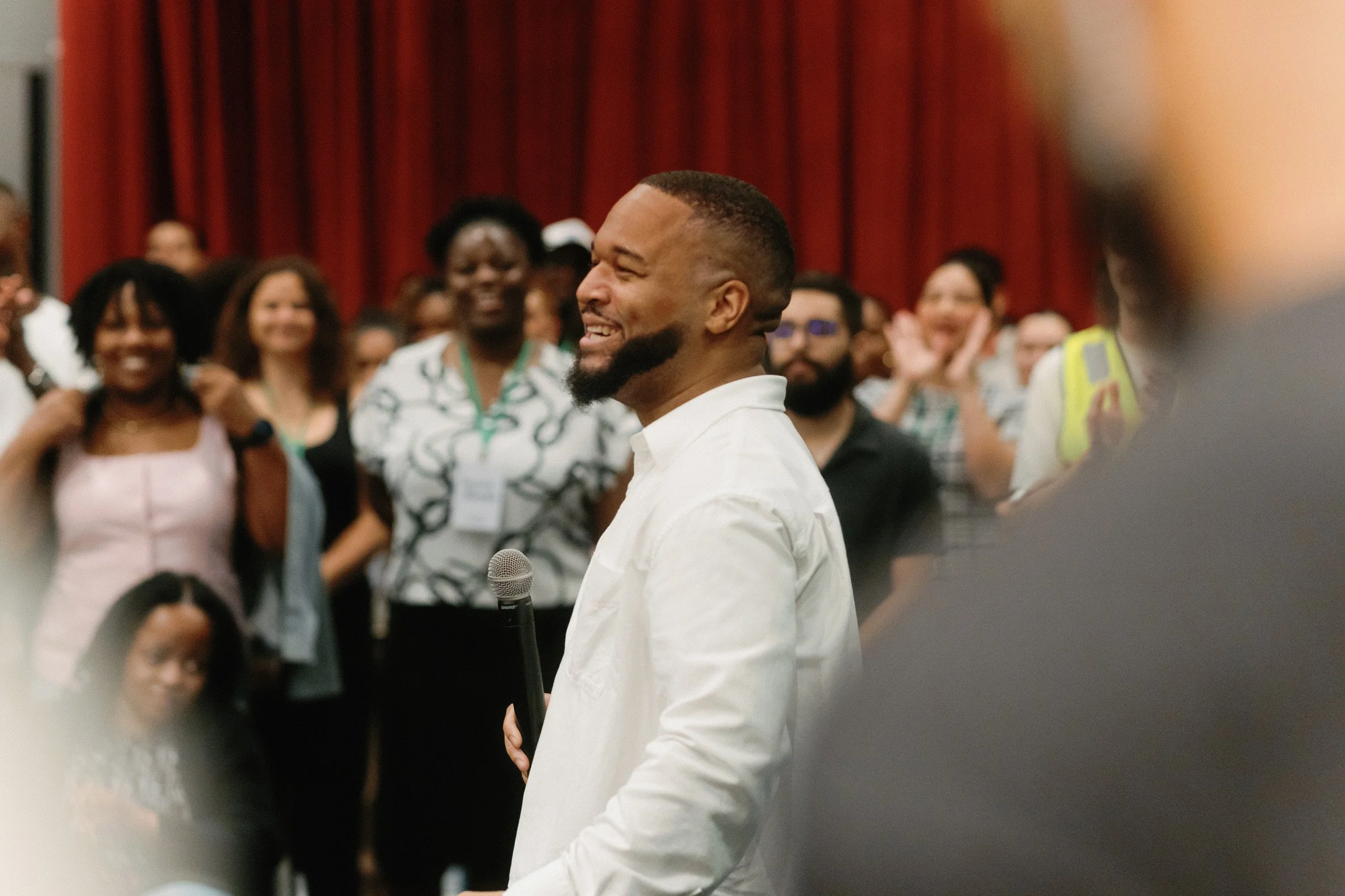 A man with a beard and mustache in a white shirt is smiling while holding a microphone. He is surrounded by an audience of diverse people who are smiling and clapping, with red curtains in the background.