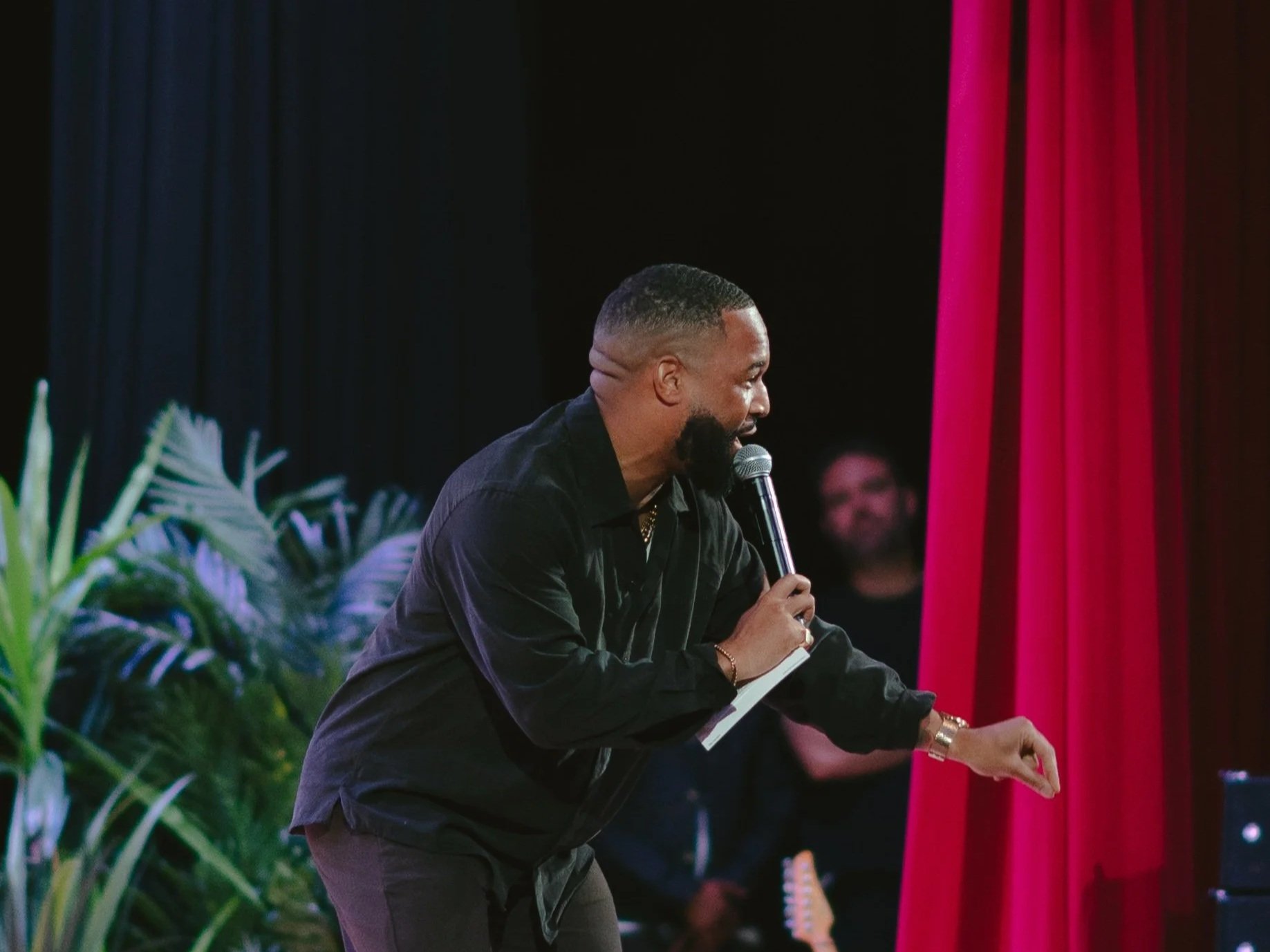 A man with short hair and a beard, wearing a black shirt, is holding a microphone and speaking at an event. There are red and blue curtains in the background and green plants in the foreground.