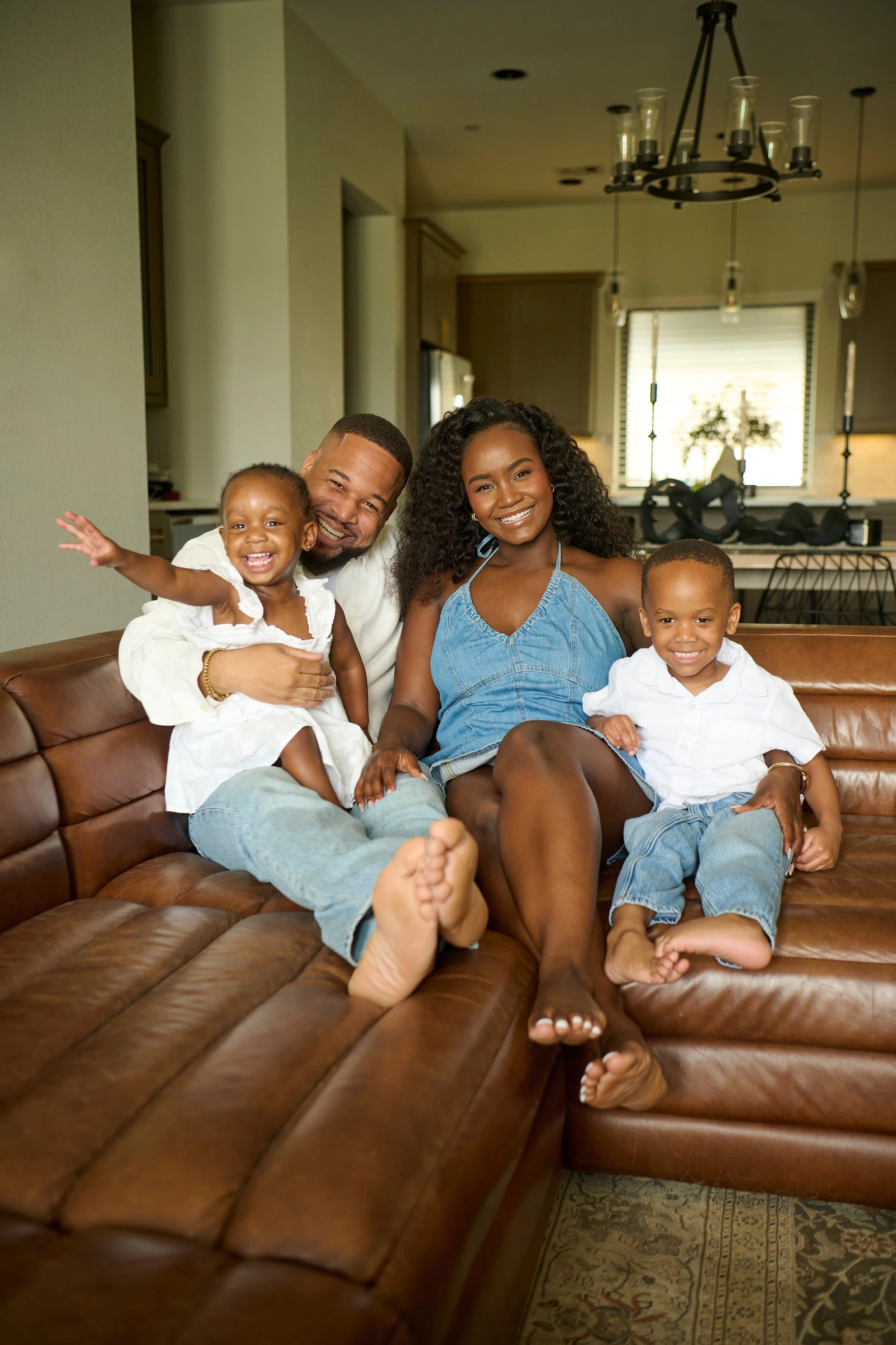 A smiling family of five sitting on a brown leather couch in a modern kitchen. The family includes a man, a woman, two young boys, and a girl. The young girl is reaching out with her arm, and the man is holding her. The woman has curly hair and wears a blue dress. The boys are in white shirts and jeans, sitting on the couch with their feet on the floor. The background features kitchen cabinets, a window with blinds, and modern pendant lights.