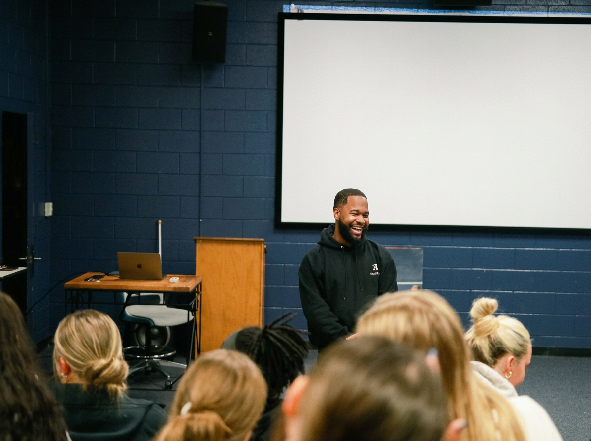 A man with a beard and black hoodie smiling and talking in front of a room of seated people, with a large blank screen behind him.