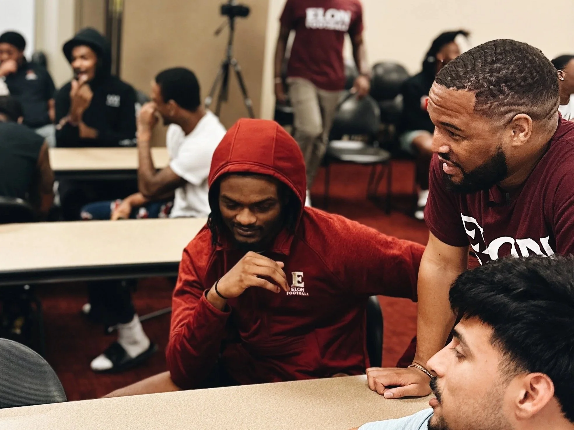 A group of young men in a classroom setting, some wearing clothing with 'Elon Football' logos, engaging in conversation and smiling. The background shows a tripod and other students, creating a lively atmosphere.