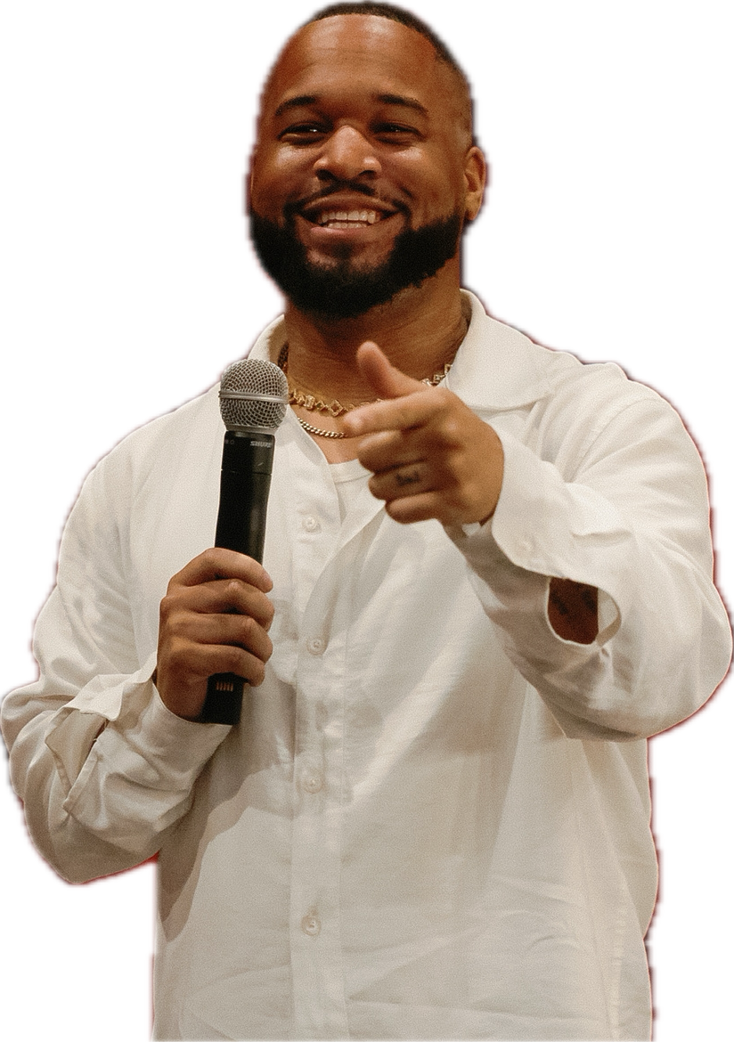A man smiling, holding a microphone in his right hand, and pointing towards the camera with his left hand, dressed in a white button-up shirt, wearing a gold chain, with a black background.
