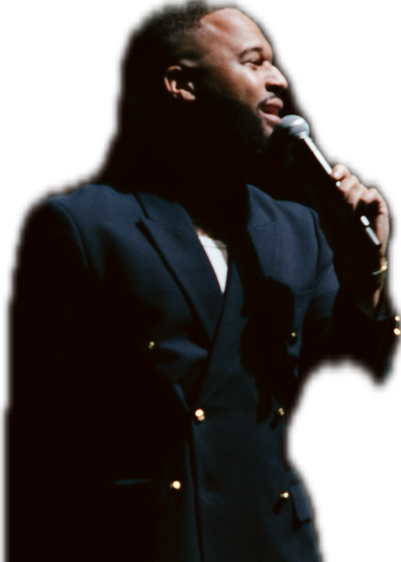 A man with dark skin and short hair is holding a microphone and appears to be speaking or singing on stage, wearing a dark blazer.