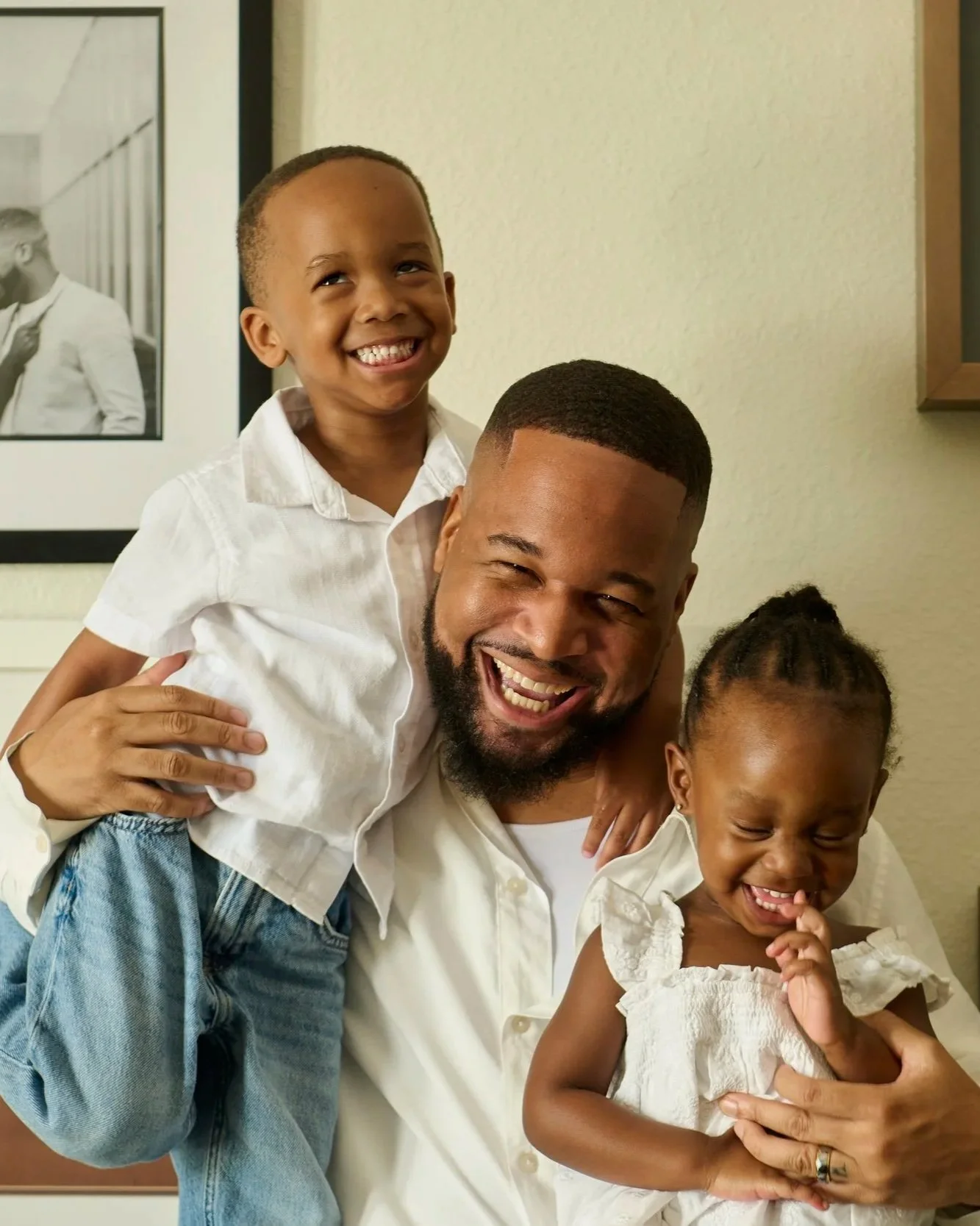 A happy family of four, with a man, two young children, and a woman, all smiling and wearing white shirts, standing indoors.