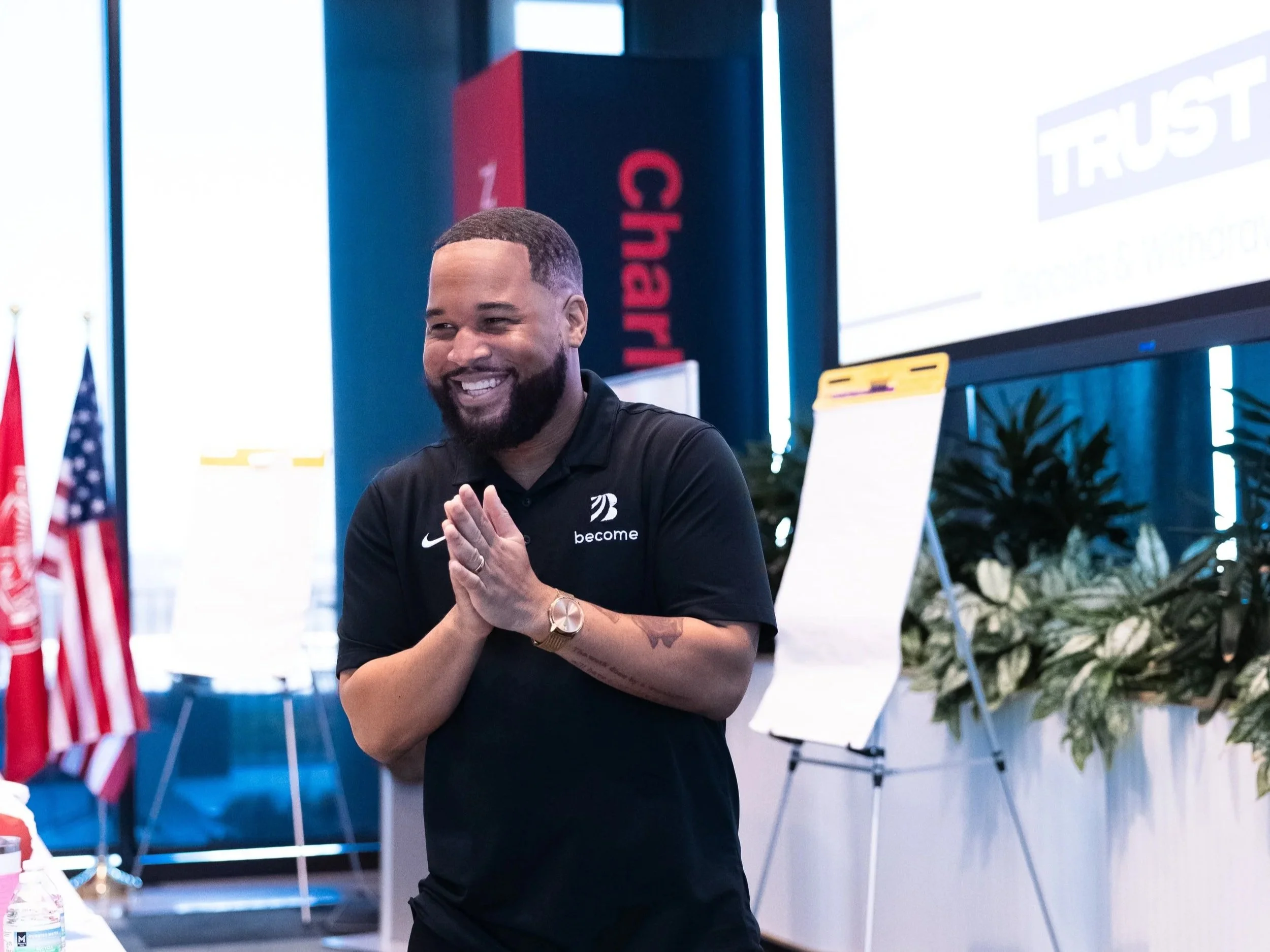 A smiling man with a beard stands indoors at a presentation or event. He is wearing a black shirt with logos and has his hands together in front of him. There are flags, a whiteboard, and greenery in the background.