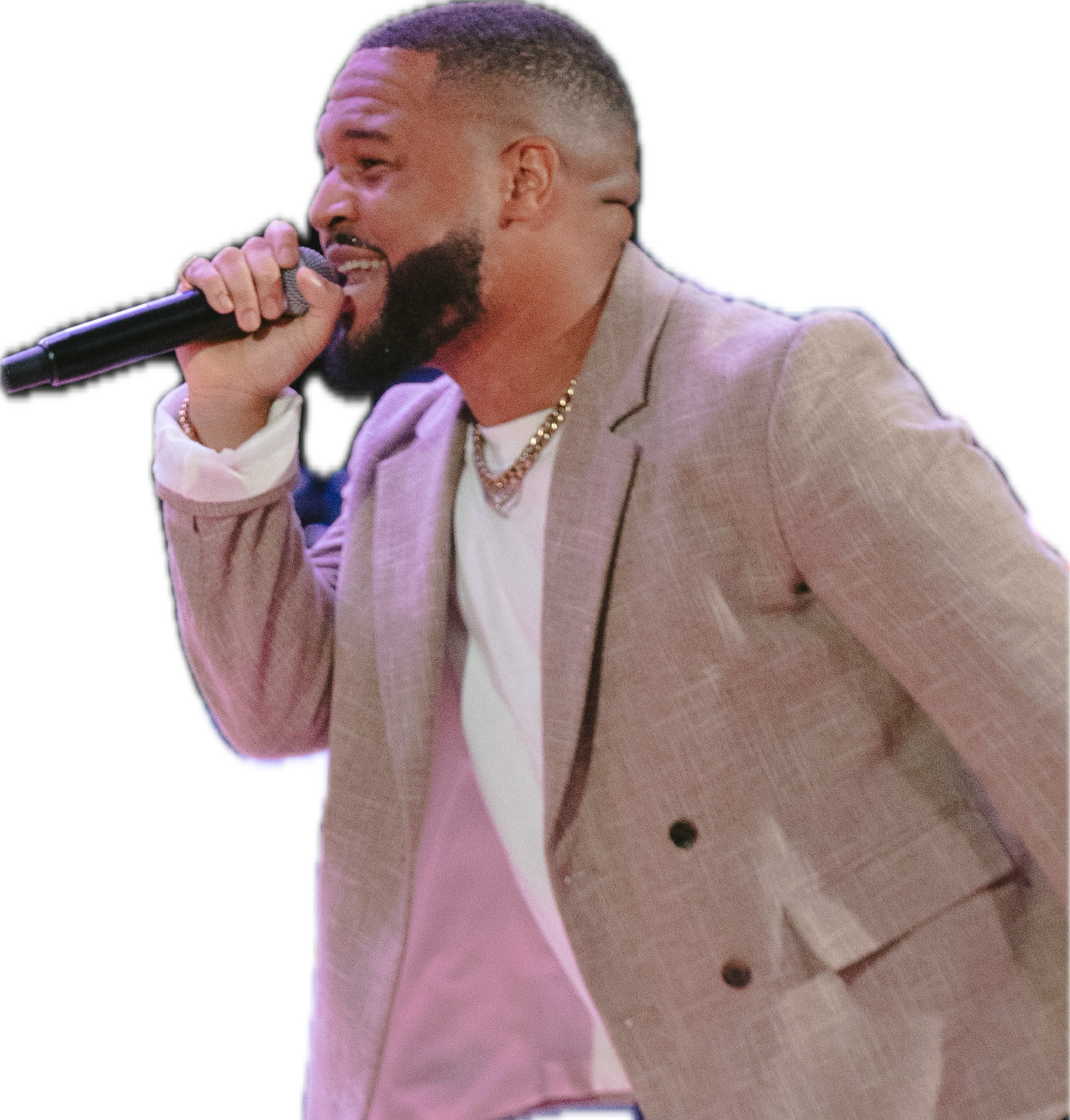 Man with short hair and beard, wearing a beige blazer and gold chain, singing into a microphone, standing on a wooden floor.