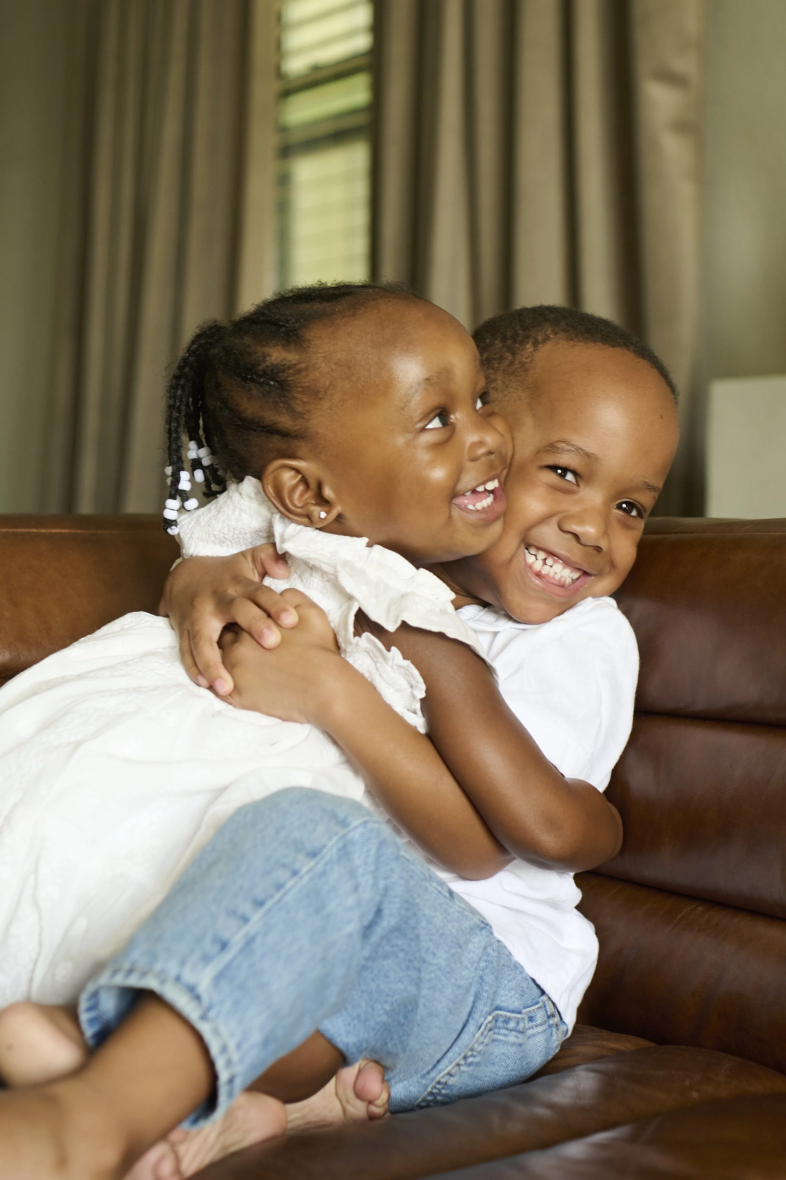 Two young children, a girl and a boy, hugging and smiling on a brown leather couch in a room with beige curtains and green window blinds.