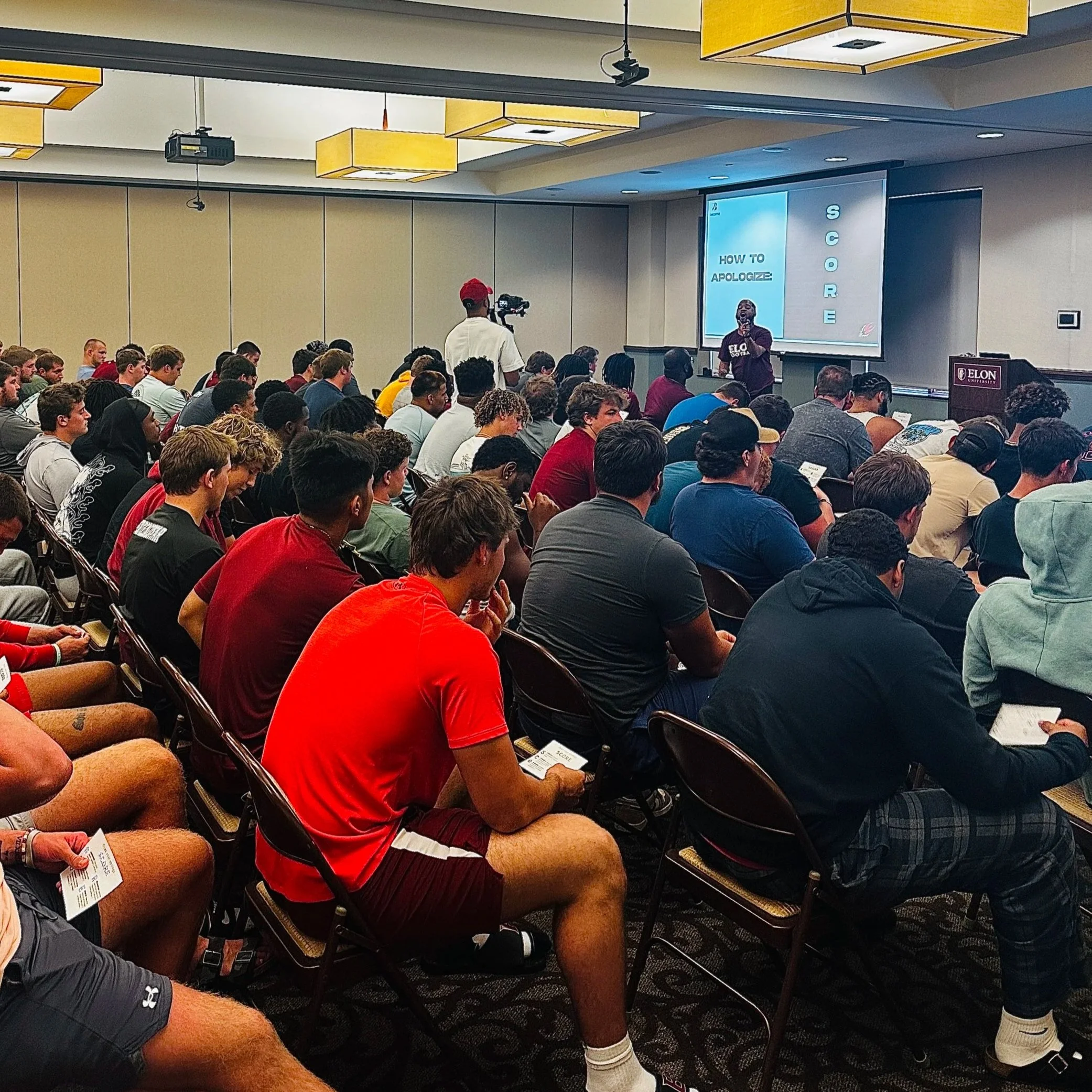 A large group of students seated in a conference room, attending a presentation. The presenter is standing in front of a screen that displays the words 'How to Apologize' and a photo of a person speaking. The room has yellow and white ceiling lights, and there is a camera or projector hanging from the ceiling.