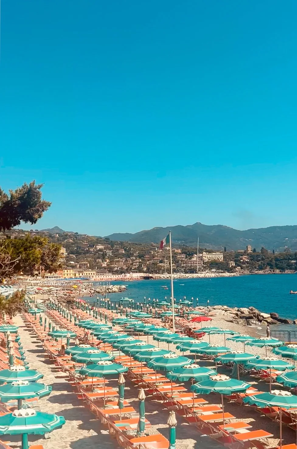 A scenic view of a beach with rows of turquoise umbrellas and orange lounge chairs, a flagpole with the Italian flag, blue water, mountains in the background, and a clear blue sky.