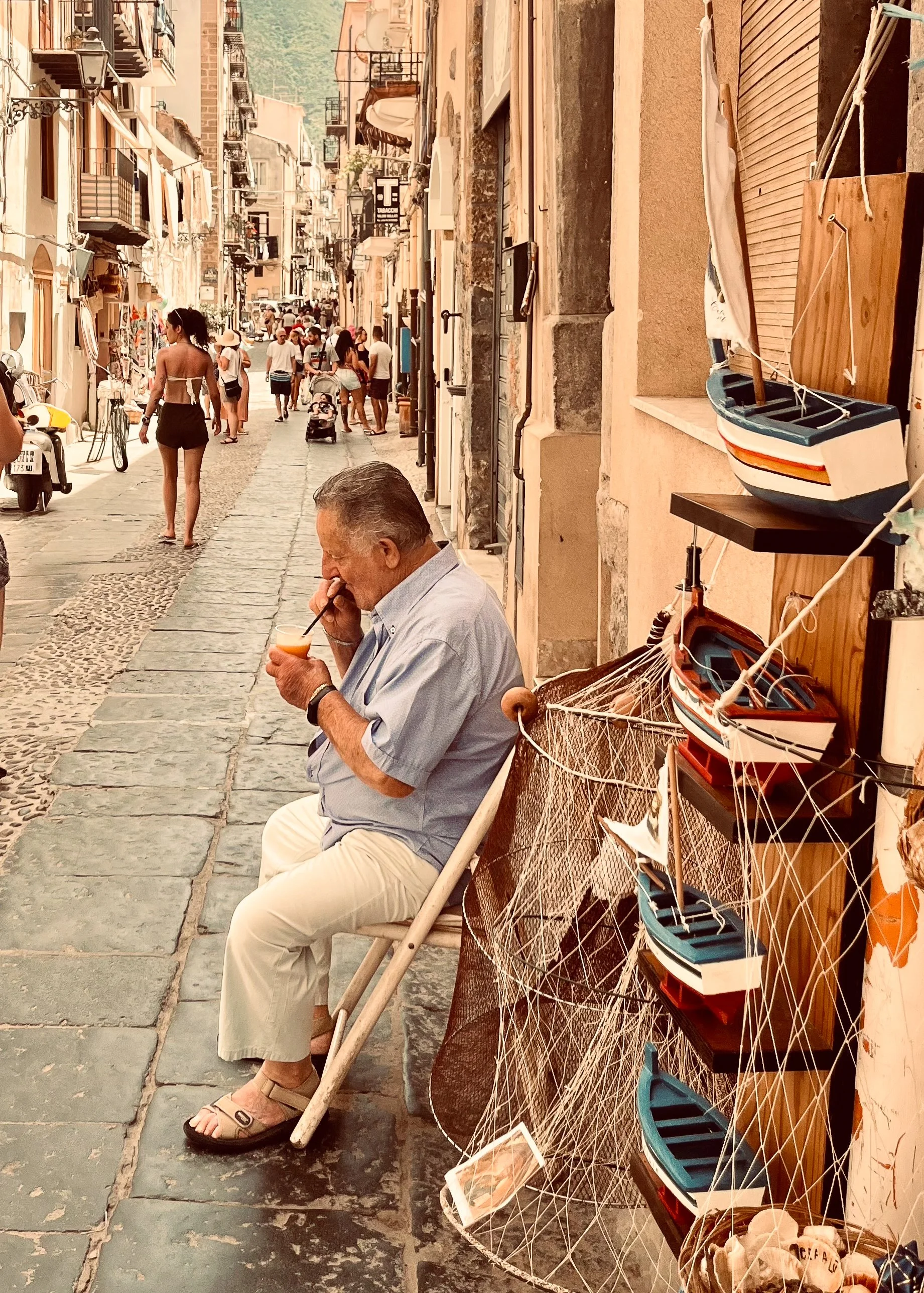 A shopkeeper takes a break on cobblestone streets of Cefalu, Sicily. Nearby, there is a display of wooden boats and fishing nets. Cefalu is one of the best places for a seaside vacation in Sicily.