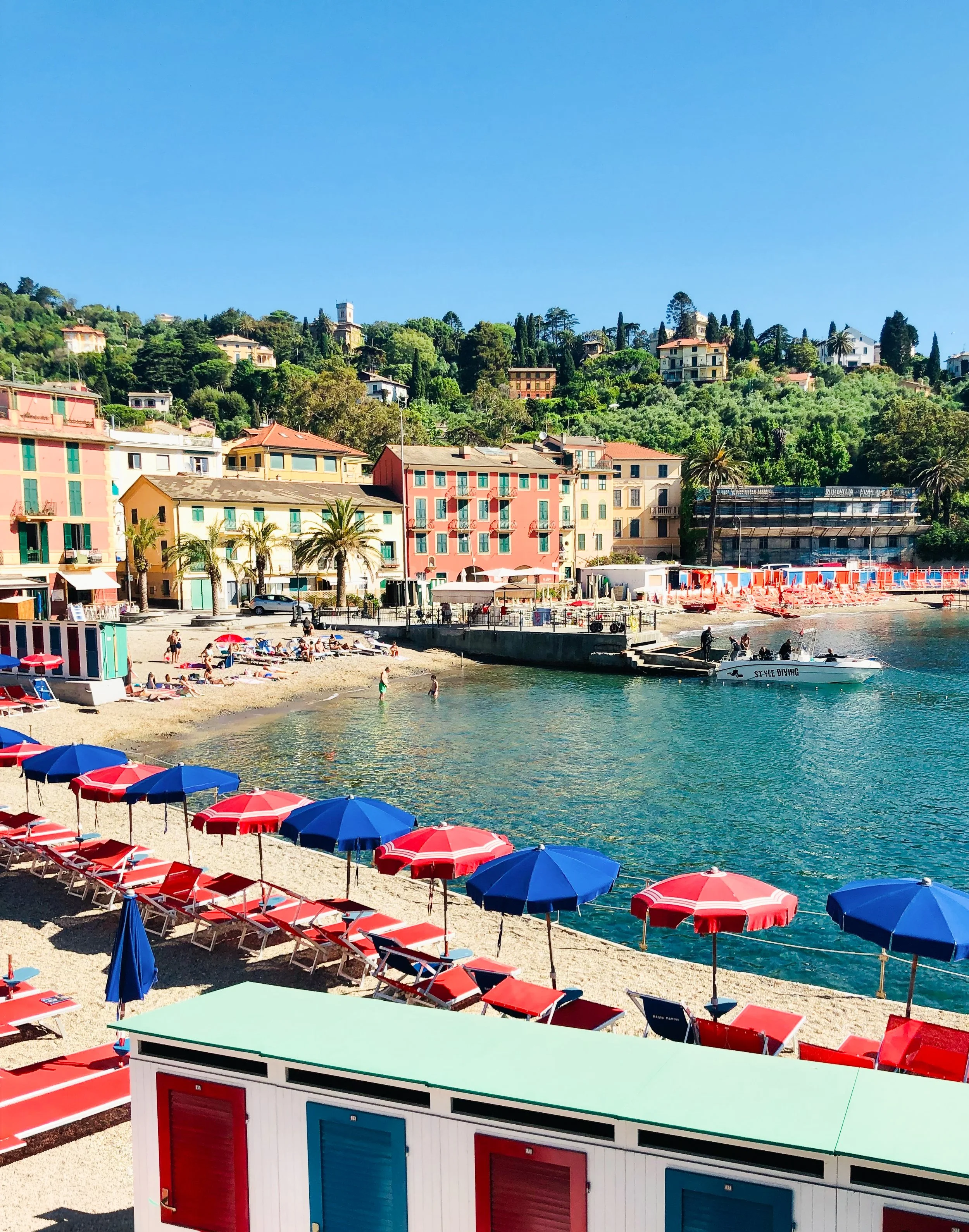 San Michele di Pagana near Rapallo, Liguria, with its colorful beachside scene of red and blue umbrellas, a small boats, a promenade, pastel-colored buildings, and lush green hillside.