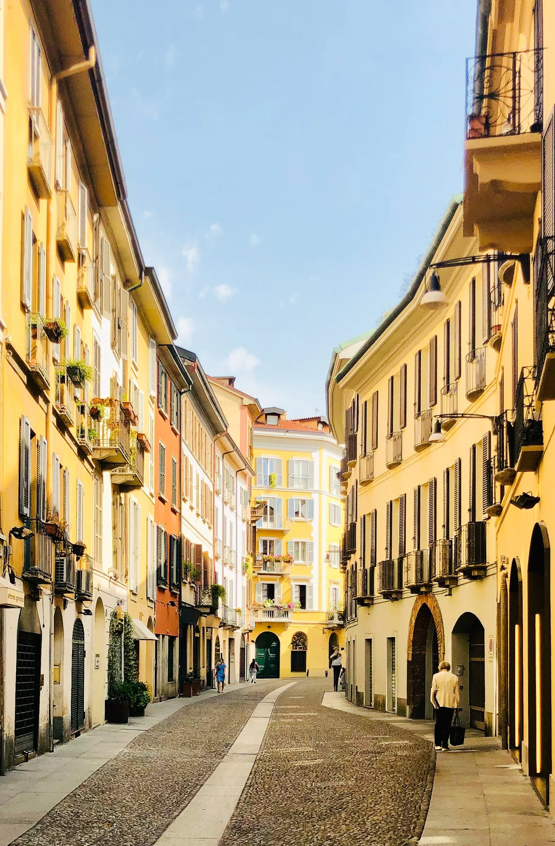 A narrow European street with colorful buildings on either side, some with balconies and flower boxes, walking pedestrians, and a blue sky overhead.