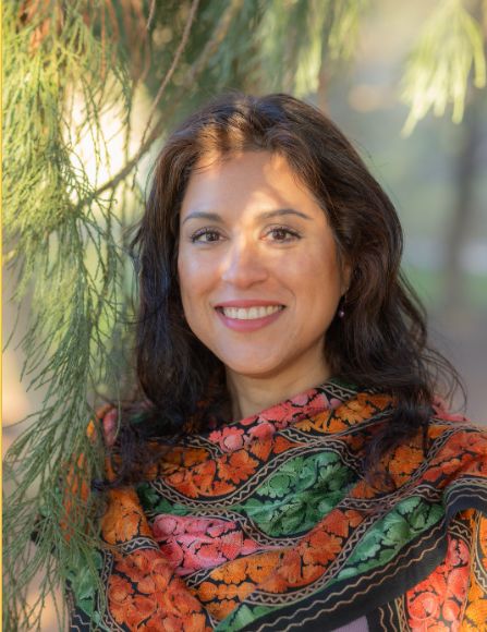 Smiling woman with dark hair outdoors, wearing a colorful floral shawl, standing near green pine branches.