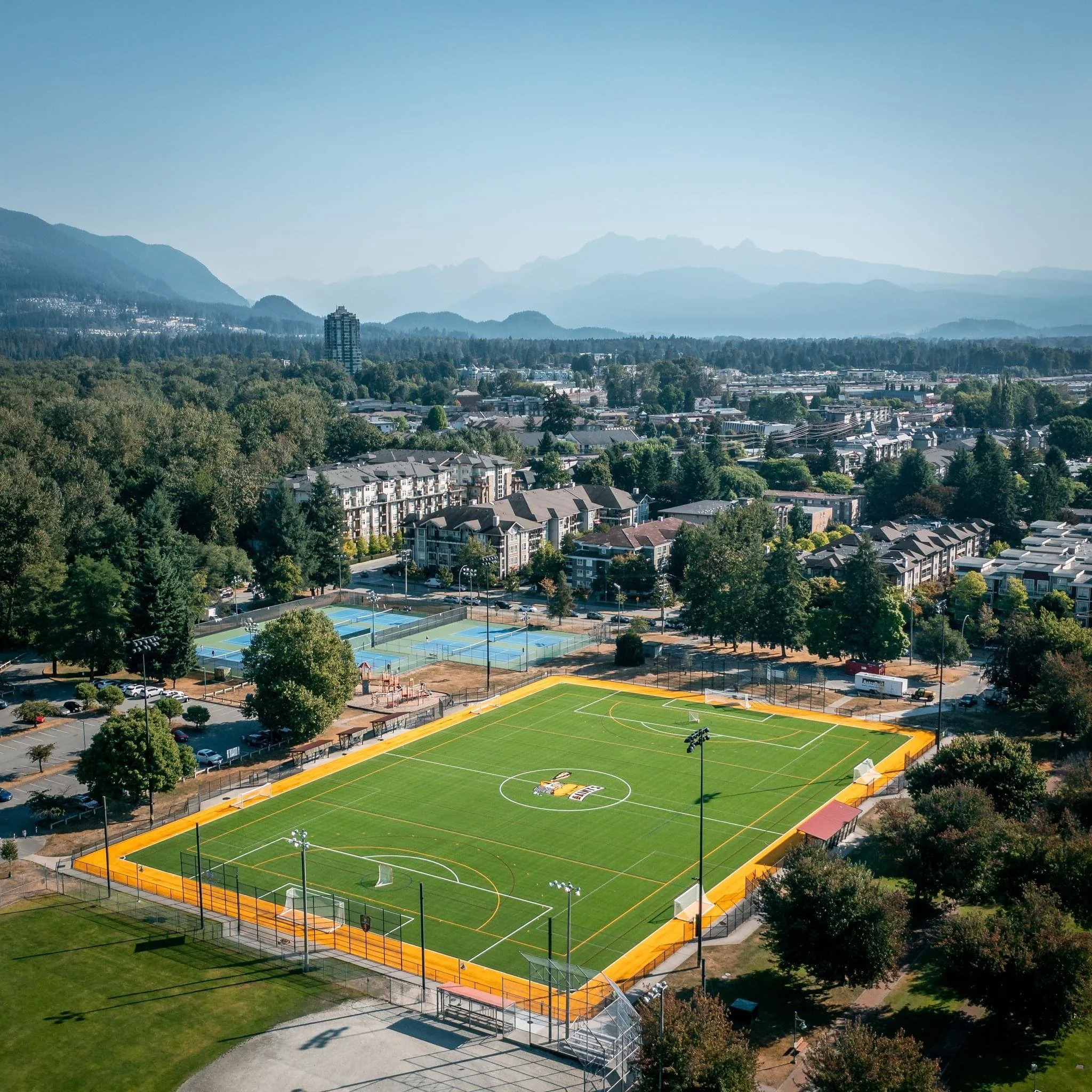 An aerial view of a sports complex with multiple tennis courts, a soccer field, and surrounding residential buildings and greenery, with mountains in the background.
