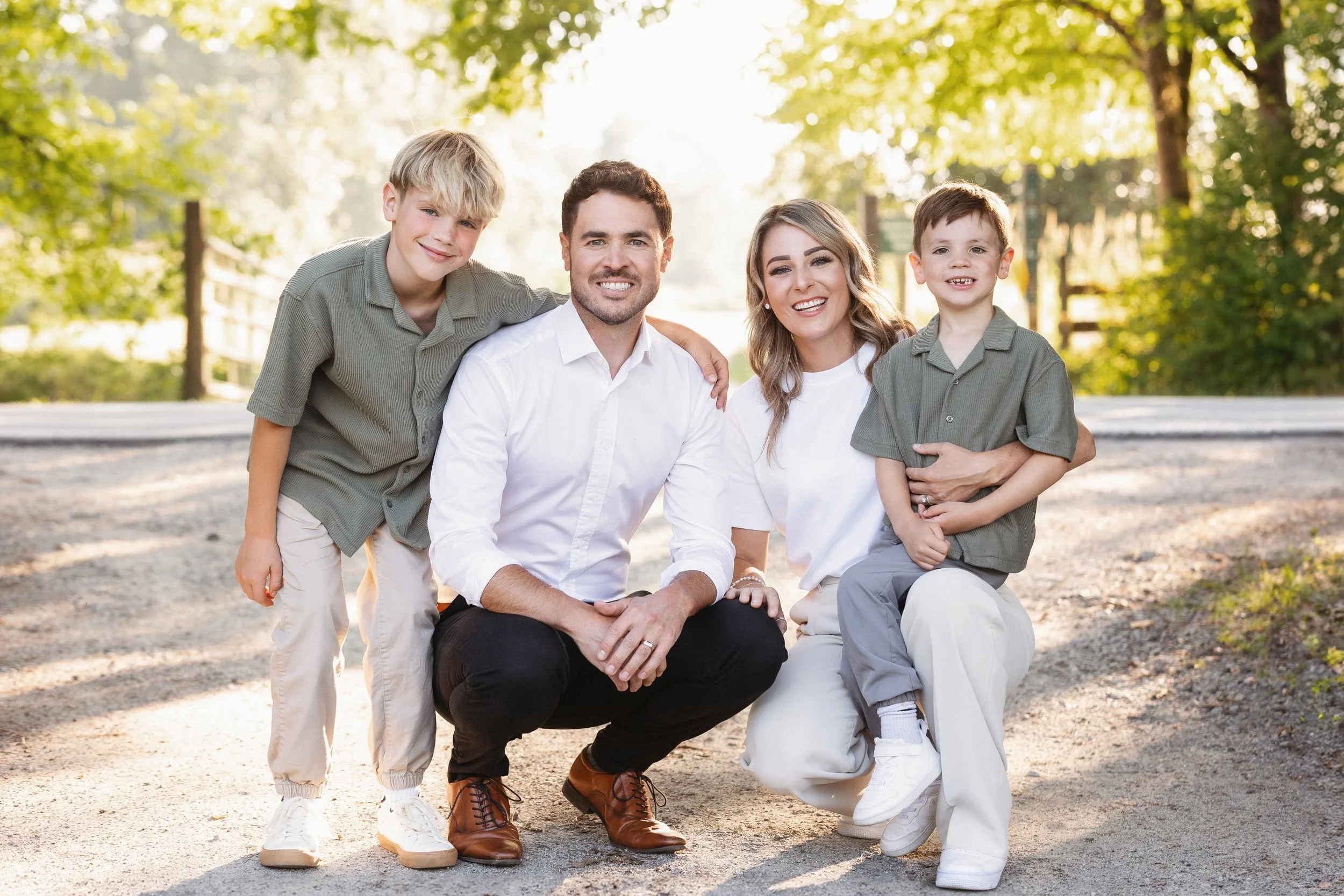 A family of four posing outdoors in a park, smiling and looking at the camera. The parents are sitting while the two children stand beside them, all with a background of green trees and sunlight.