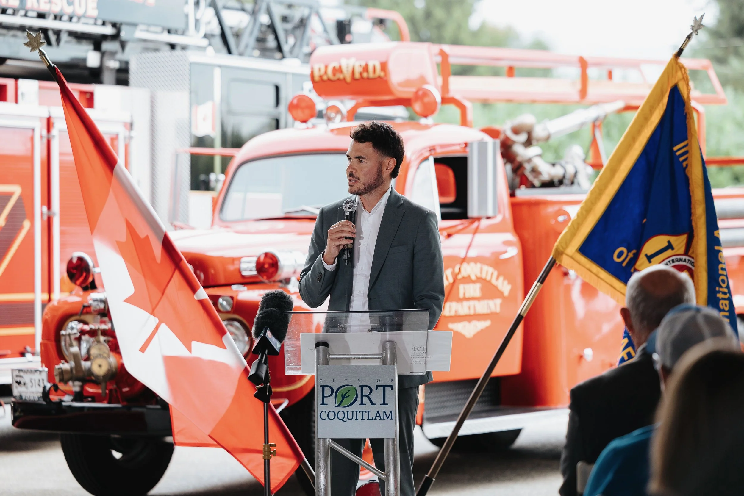 A man in a gray suit speaks into a microphone at an outdoor event with fire trucks and flags in the background. A sign on the podium reads "PORT COQUITLAM".