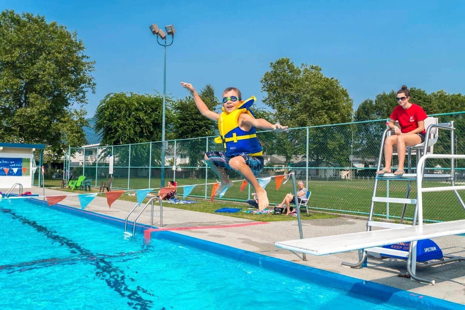 A boy in swimming gear jumping into a swimming pool with a colorful flag banner hanging above the poolside, while a girl in sunglasses sits on a lifeguard chair overseeing the area.
