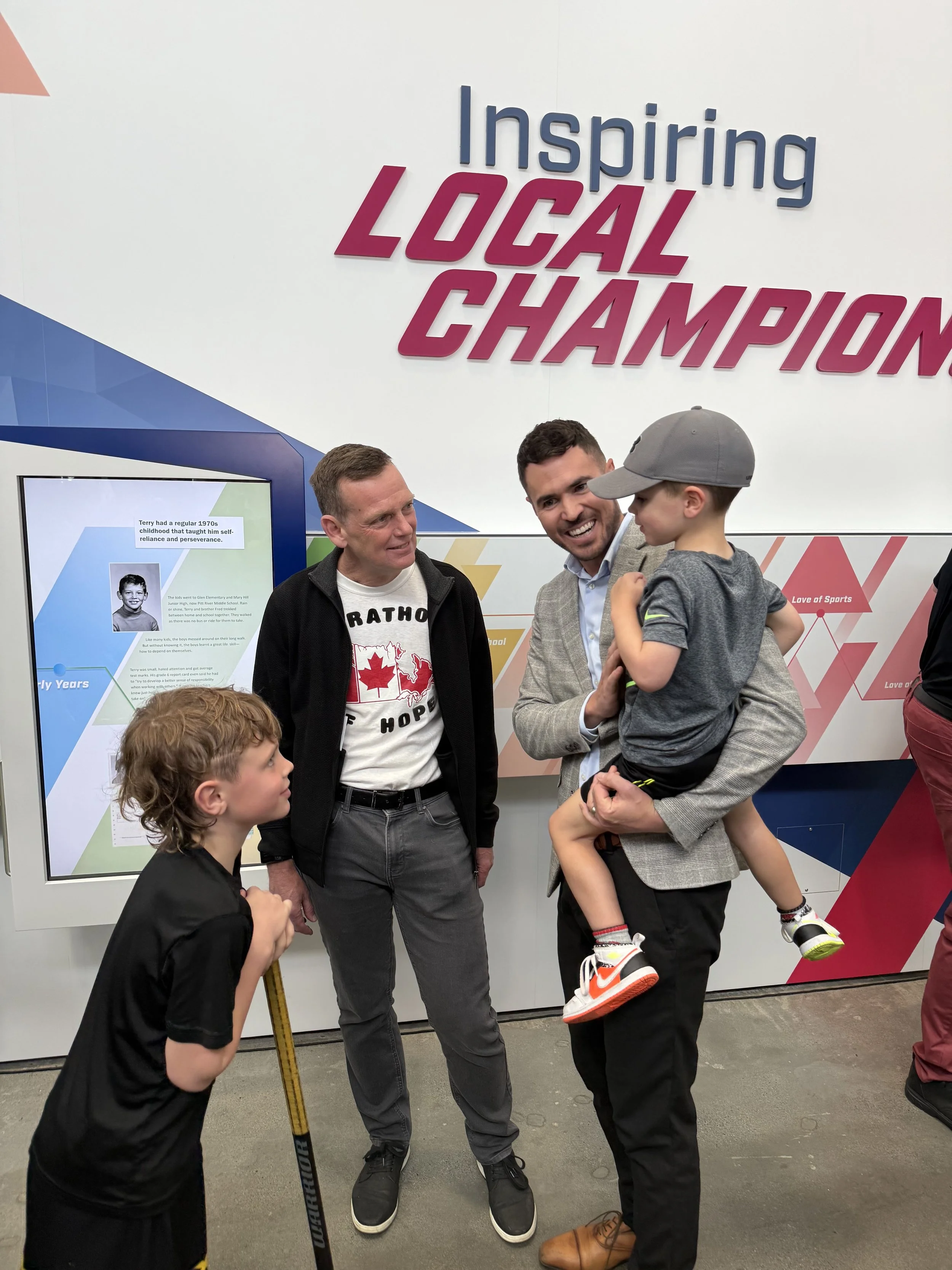 Three adults and two children are standing and engaging in conversation at an indoor event with a wall that says 'Inspiring Local Champion' in the background. One man is holding a young boy.
