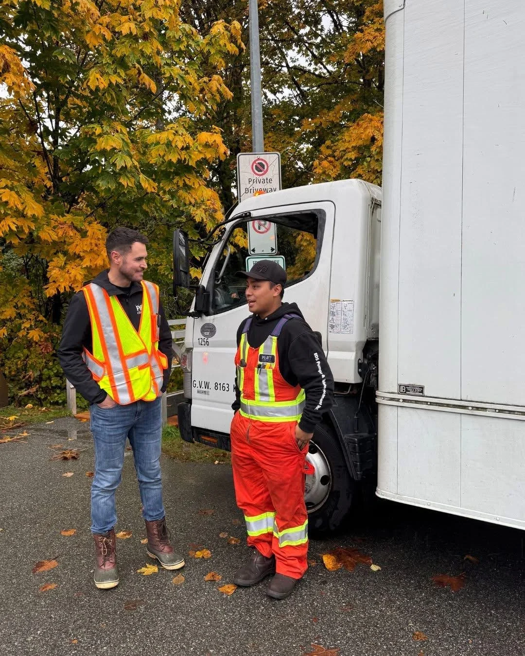 Two men in safety vests and work attire standing near a white truck on a wet pavement with fallen autumn leaves, talking and smiling, with trees in autumn colors and a no parking sign in the background.