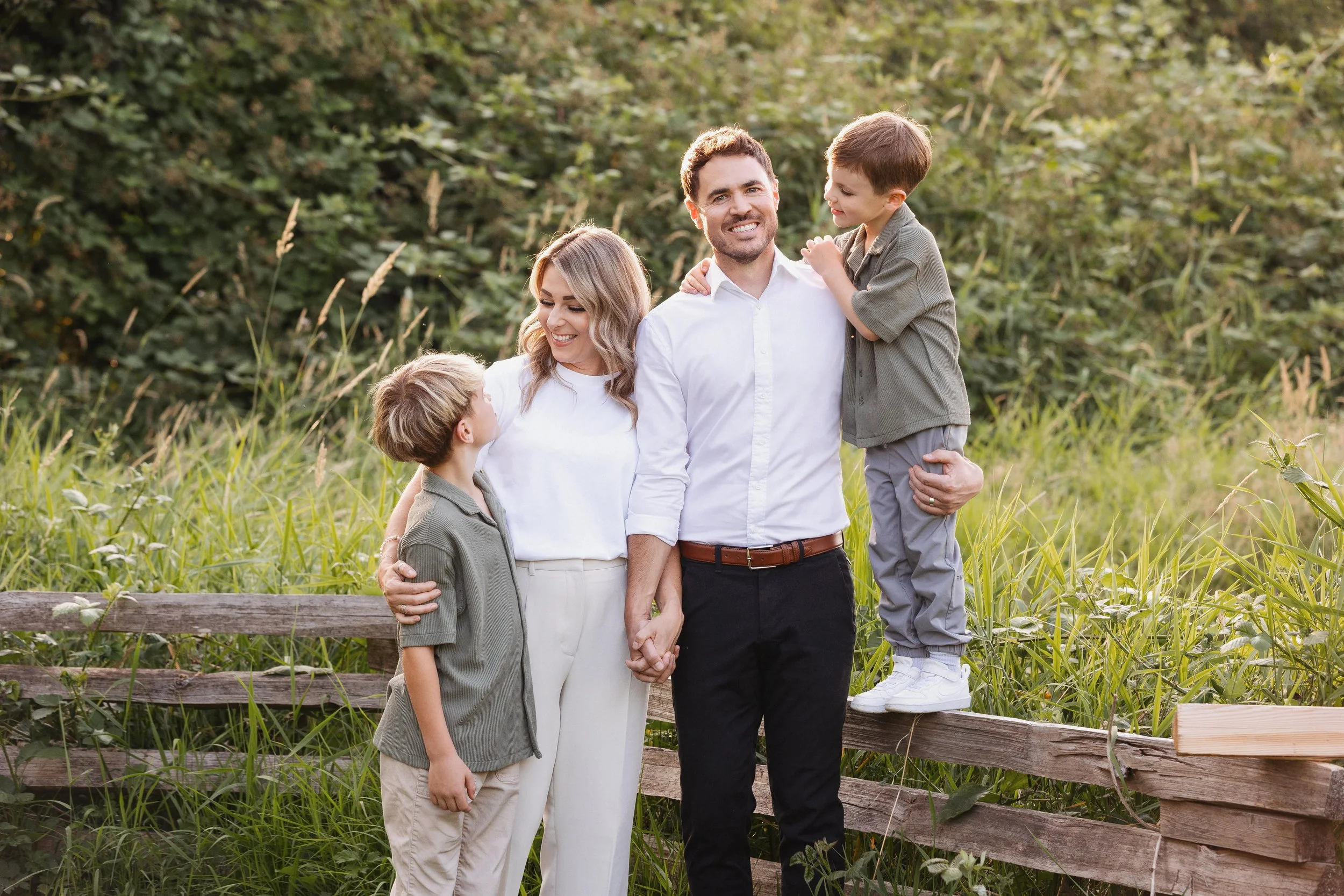 A family of four—the father, mother, and two young boys—stands outside in a grassy area with trees in the background. They are holding hands and smiling, enjoying a pleasant moment together.