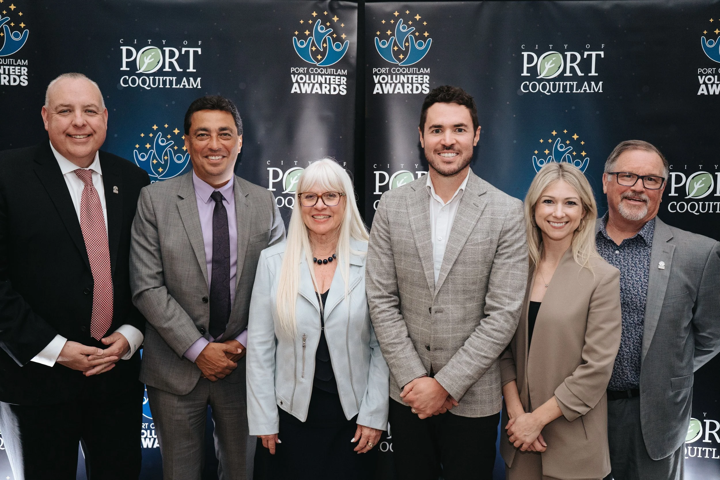 Group of six adults standing together at the Port Coquitlam Volunteer Awards event, smiling in front of a backdrop with the event logo.