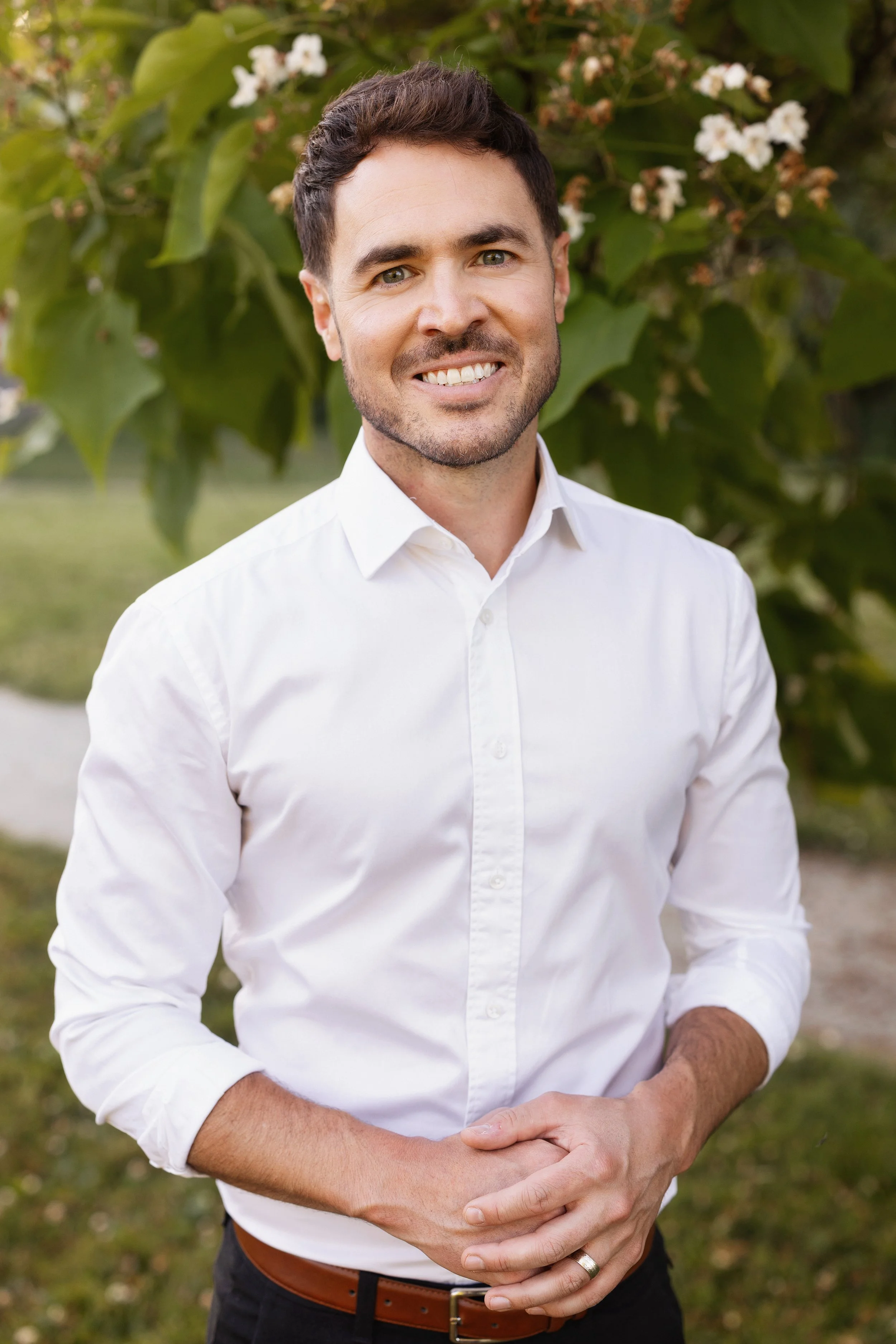 A young man with dark hair, green eyes, and light stubble, smiling and standing outdoors in front of green foliage with white flowers, wearing a white button-up shirt and black pants.