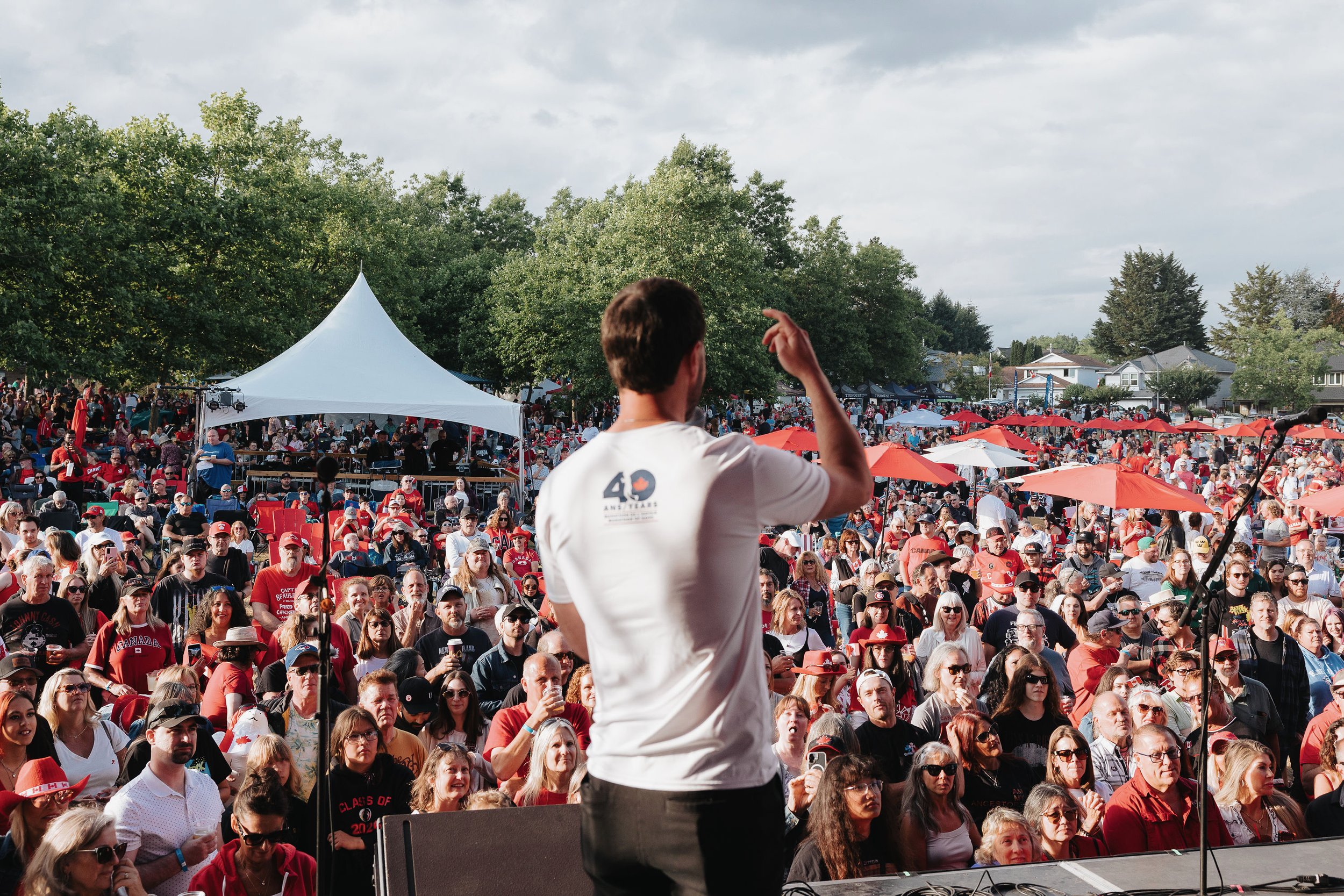 A crowd of people watching a speaker at an outdoor event with a white canopy and red umbrellas, some wearing Canadian flags.