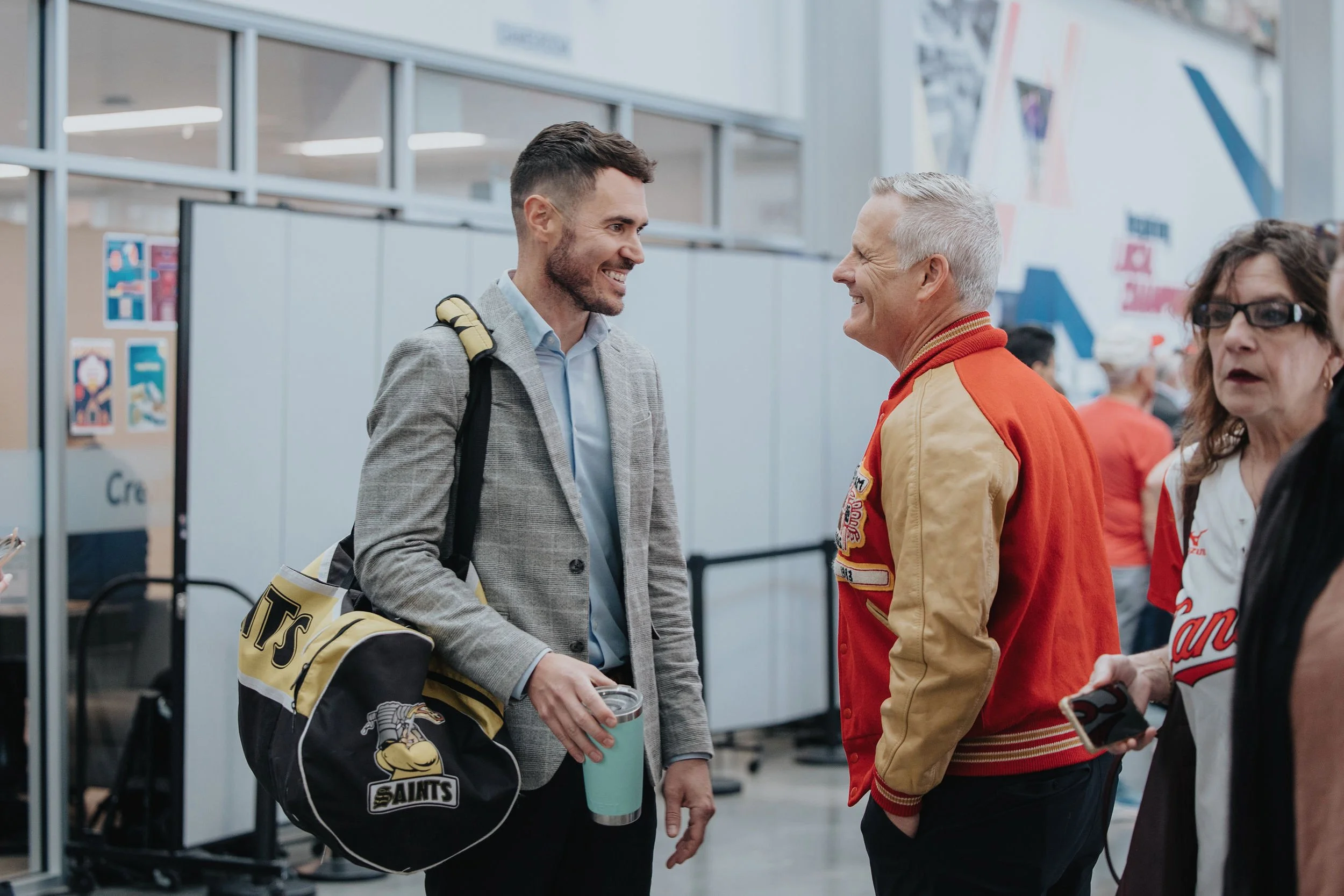 Two men smiling and talking in an airport, one with a Saints hockey bag, the other wearing a red and yellow jacket. A woman with glasses is in the background.