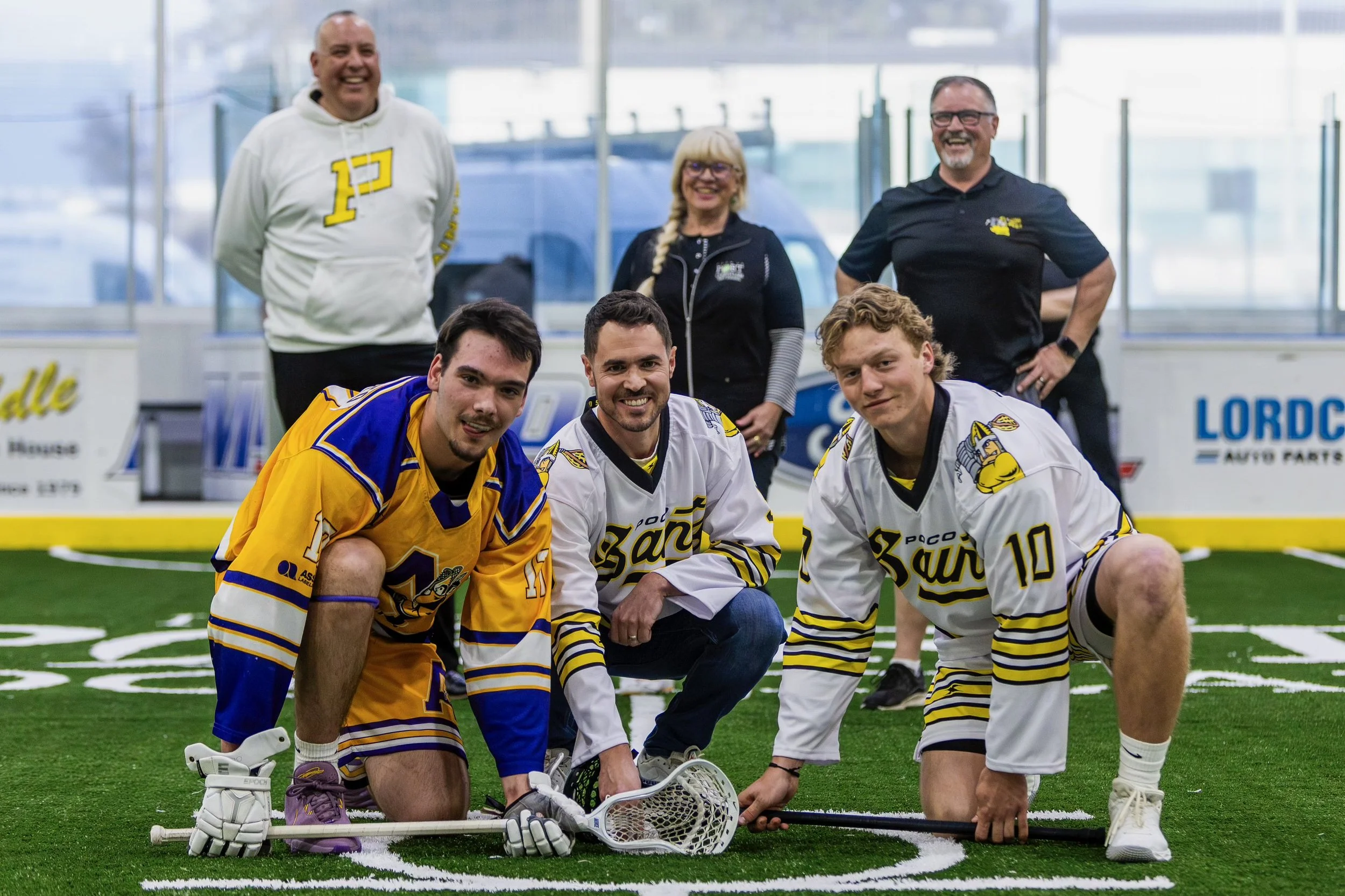 A group of men and women posing on an indoor lacrosse field, some kneeling and some standing, wearing sports jerseys with 'Pace' and yellow and blue colors, and smiling at the camera.