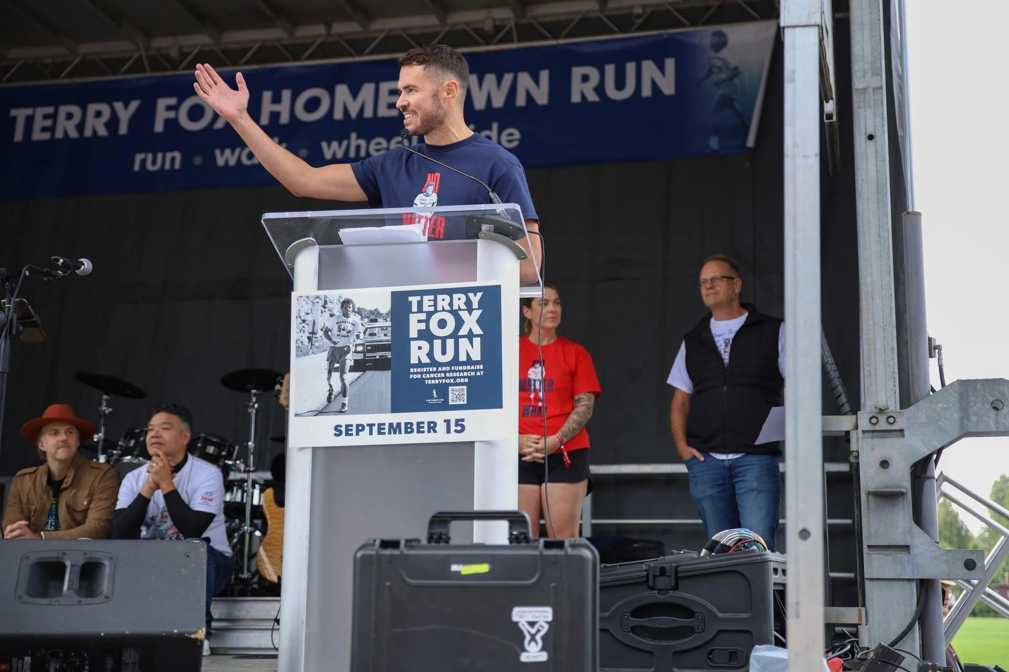A man speaking at a podium during a race event with a banner that says 'Terry Fox Run' in the background. Several people are sitting and standing nearby on stage.