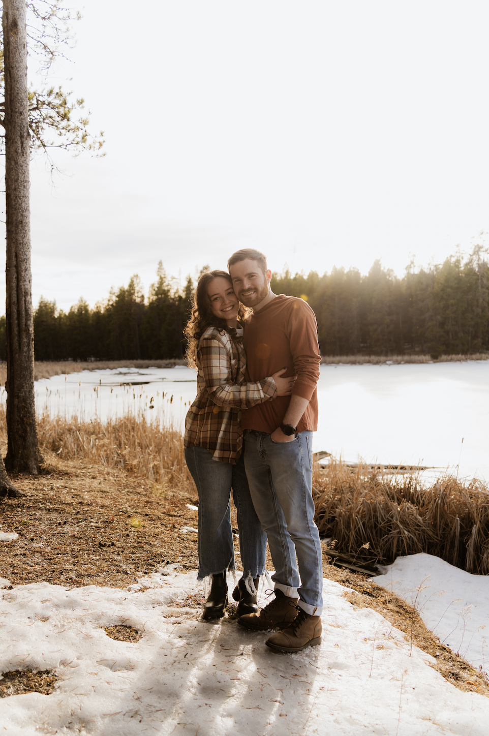 A happy couple hugging outdoors by a lake with snow and trees, during sunset.