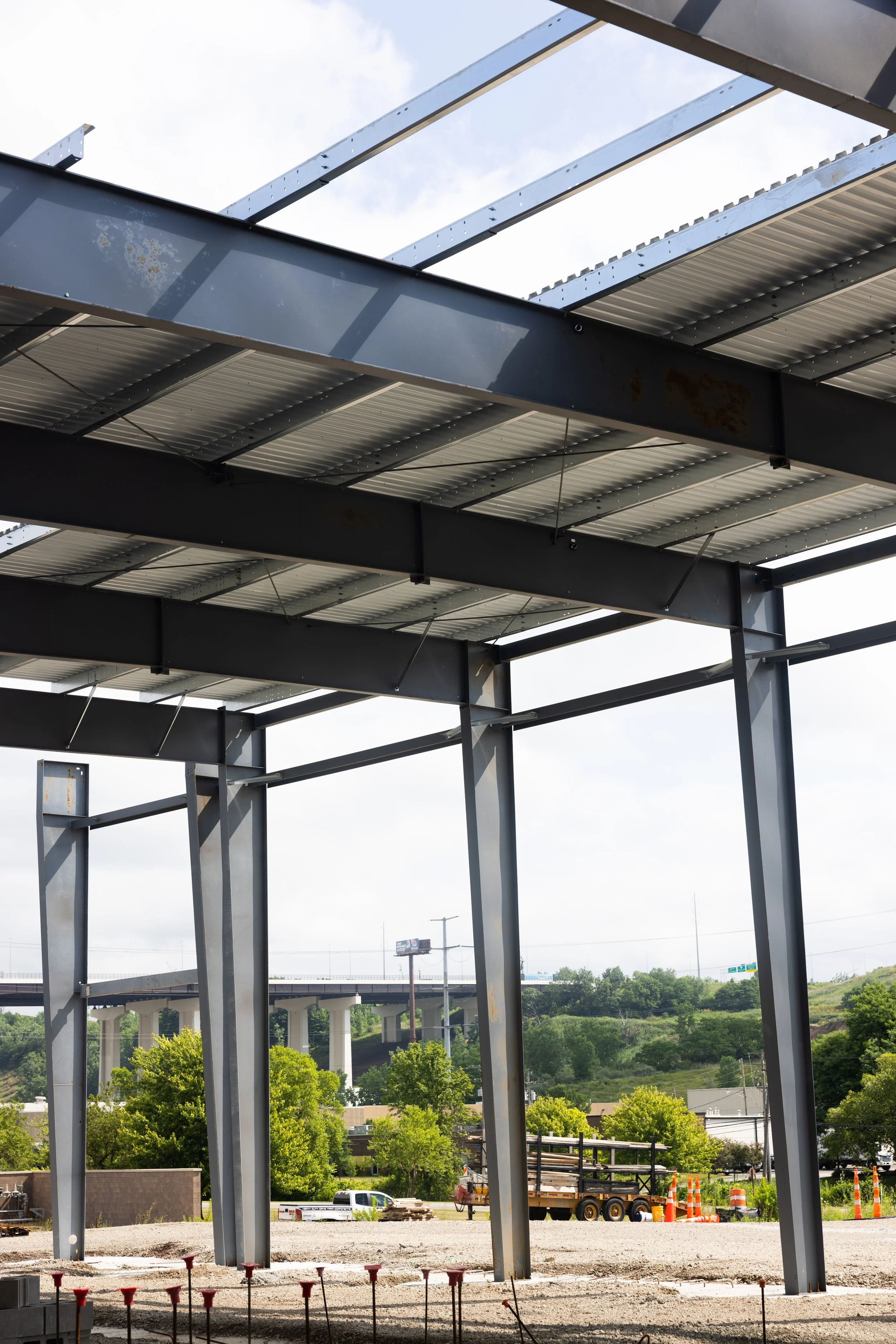 Steel framework of a building under construction with visible girders and support beams, construction site with trees and highway in the background.