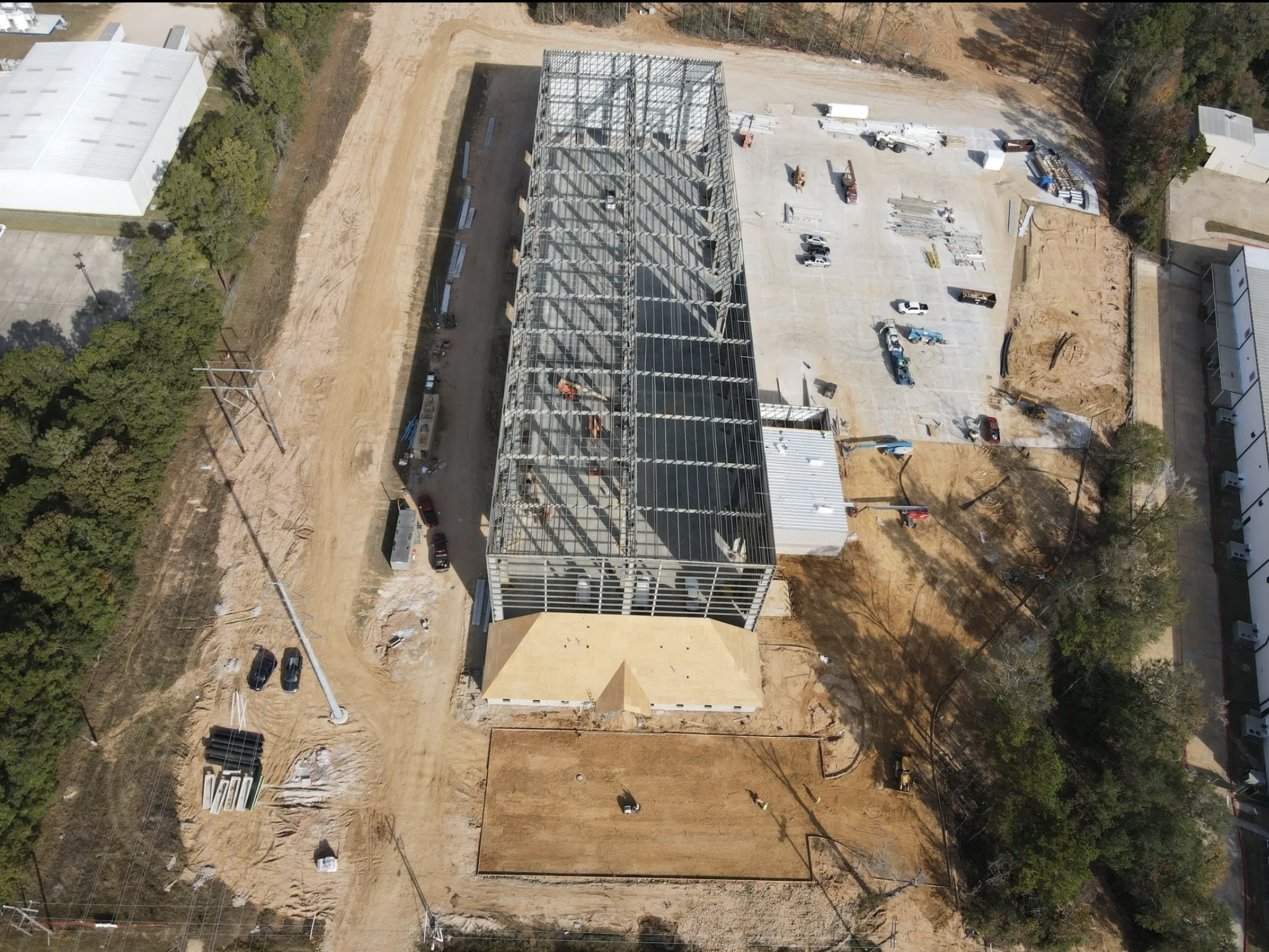 Aerial view of a building under construction with a steel framework, surrounded by construction vehicles, equipment, and partially developed land in a wooded area.